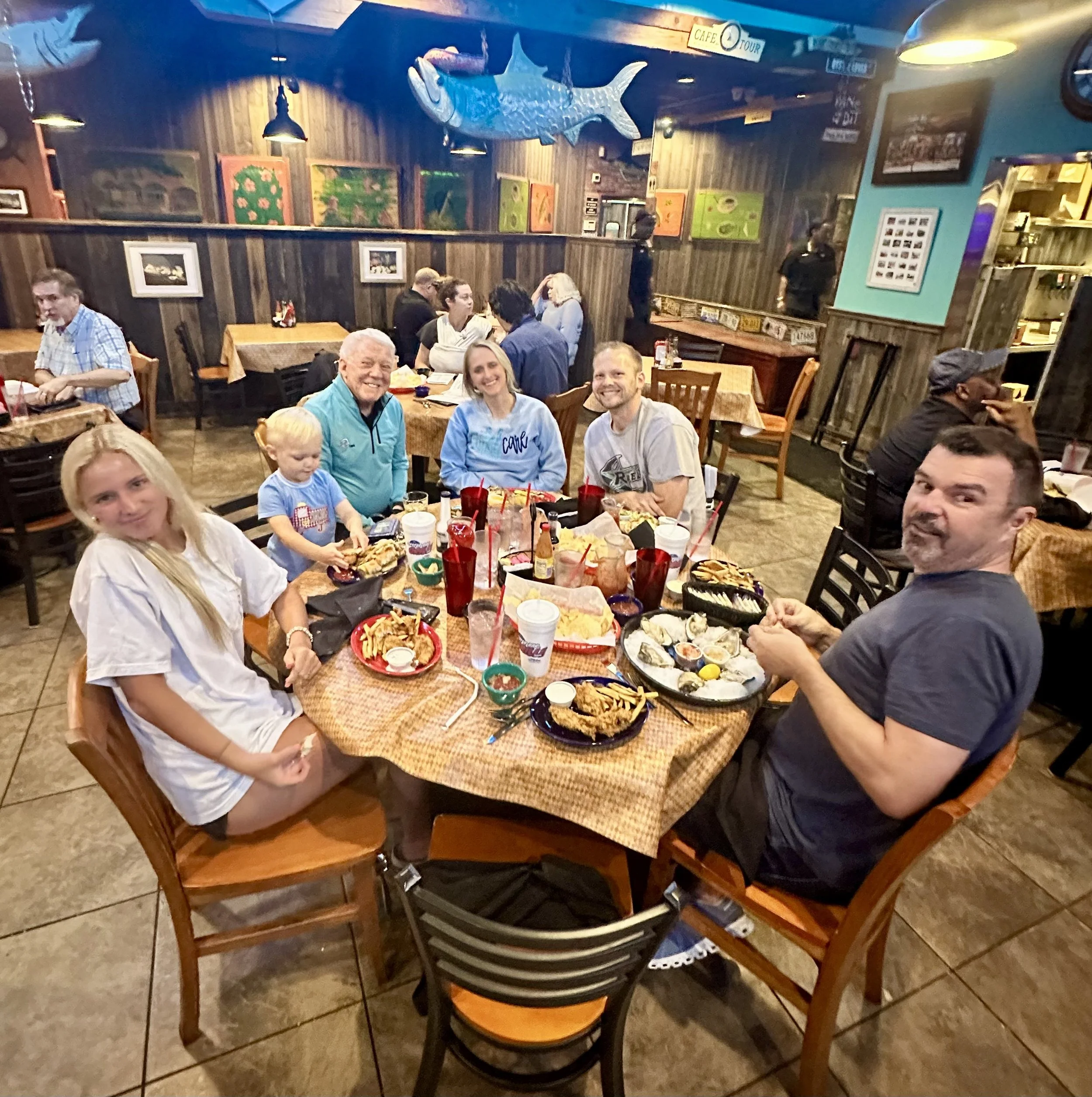 People dining in a rustic restaurant with wooden walls and fish decorations, enjoying food at tables with plates of food and drinks.