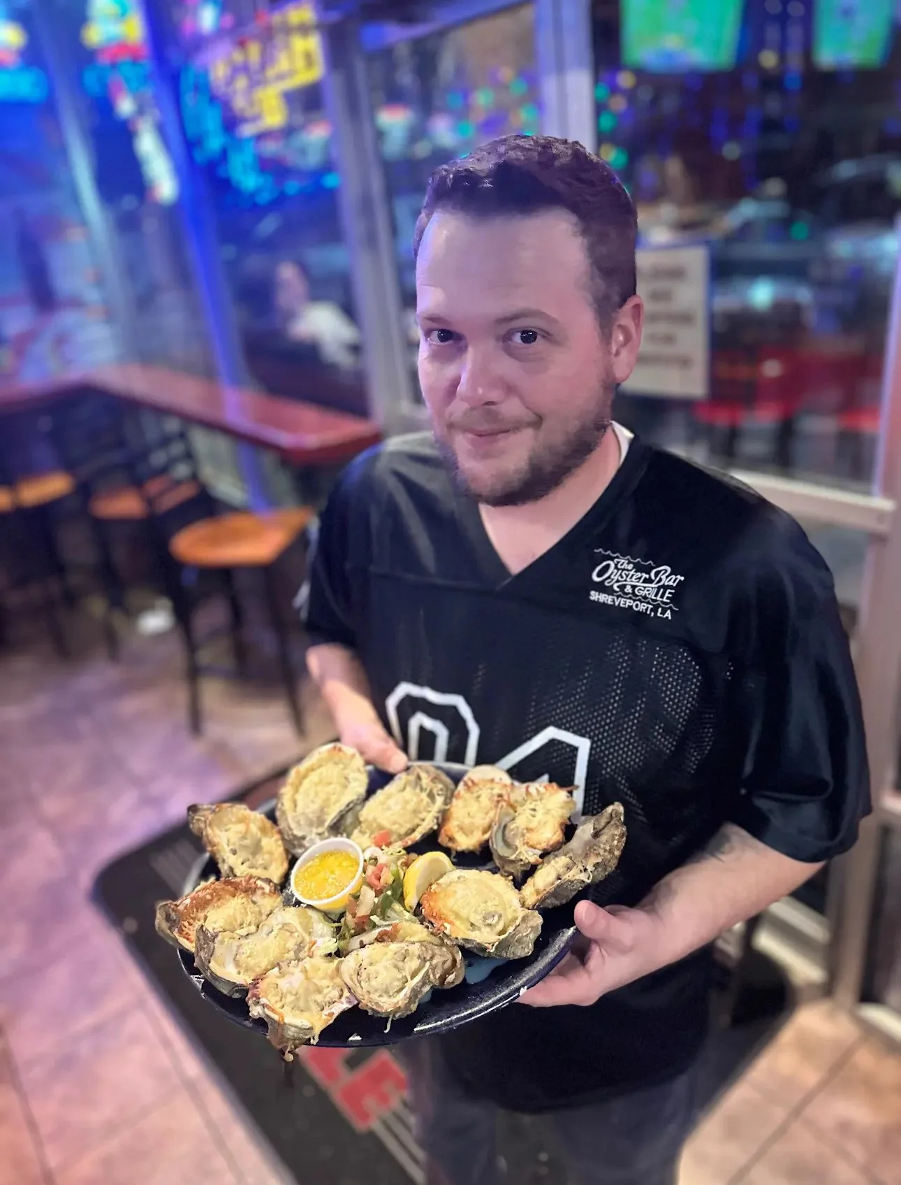 Man holding a plate of oysters at a seafood restaurant in Shreveport, Louisiana, with vibrant neon lights in the background.