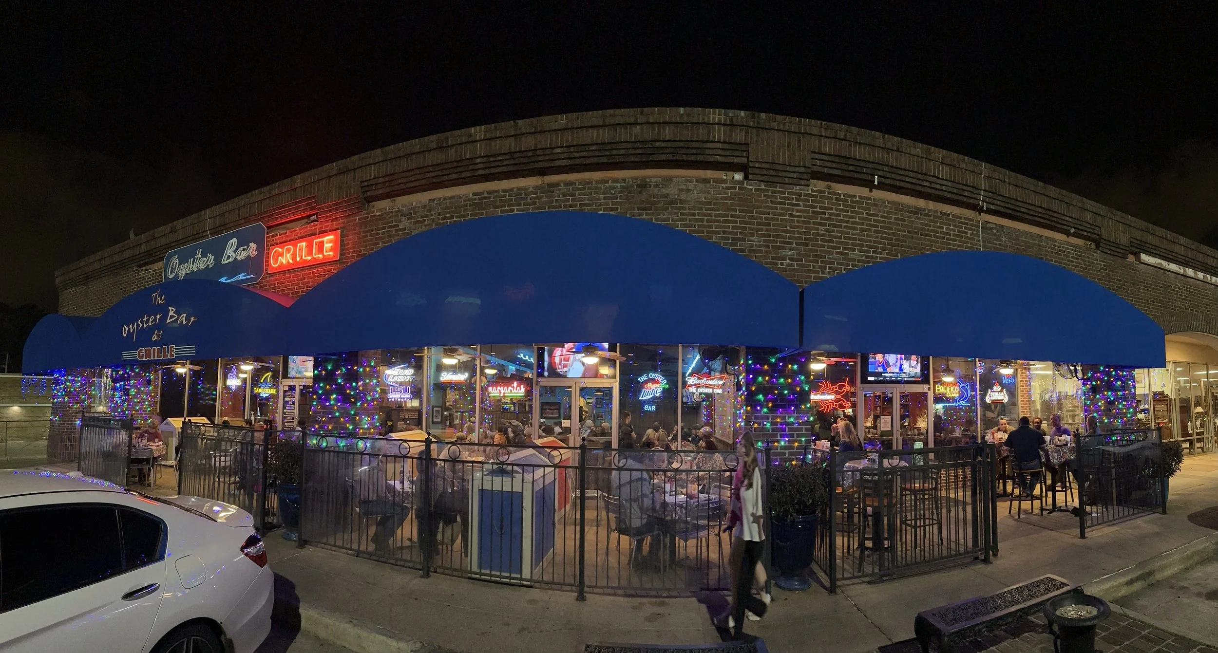 Night view of a restaurant called 'The Oyster Bar & Grill' with neon signs and colorful string lights. The restaurant has large blue awnings and indoor seating visible through large windows. People are seated indoors and walking outside.
