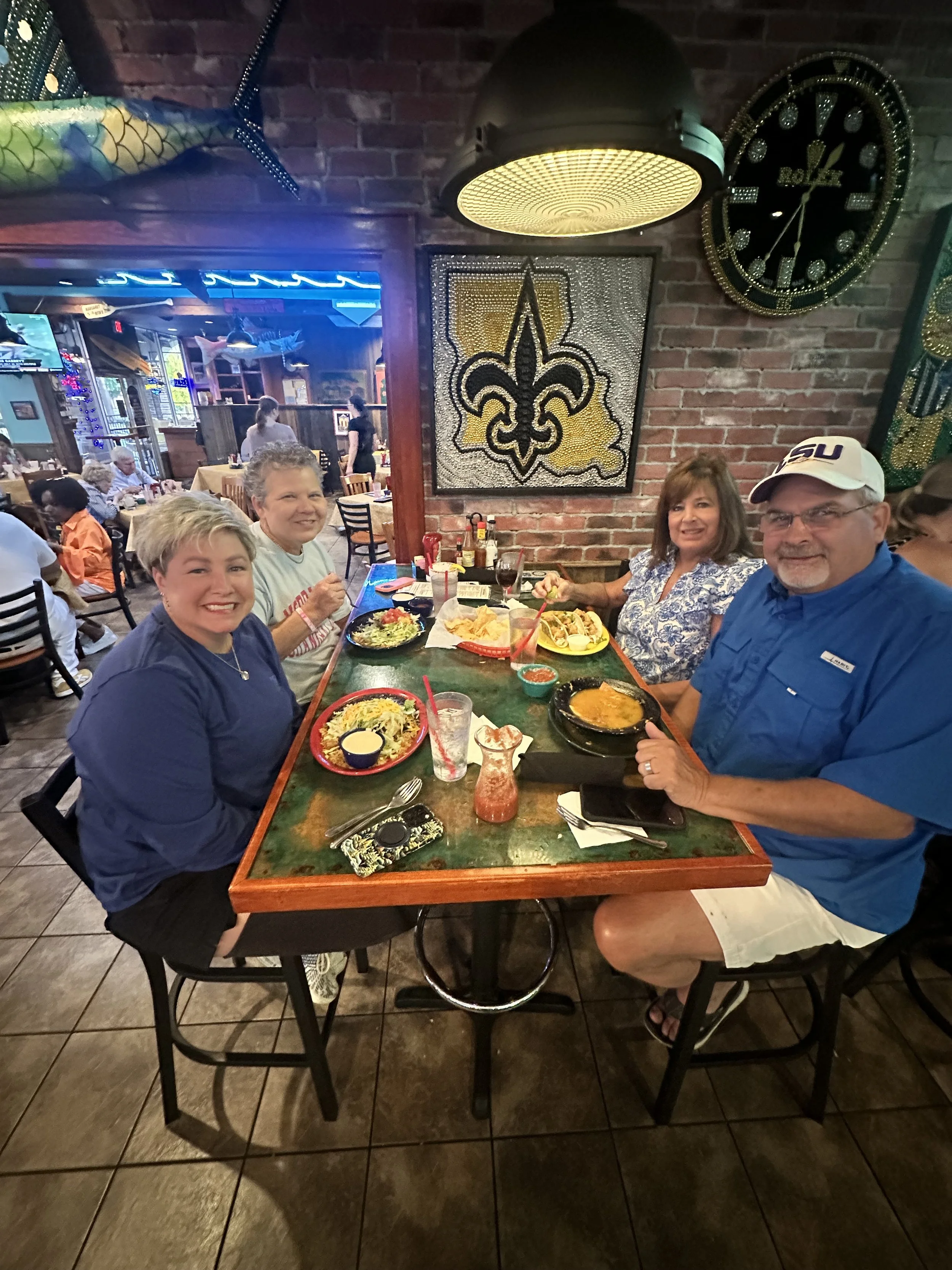 A group of four people sitting at a restaurant table with food, drinks, and condiments, smiling and posing for a photo with a brick wall and decorative art behind them.