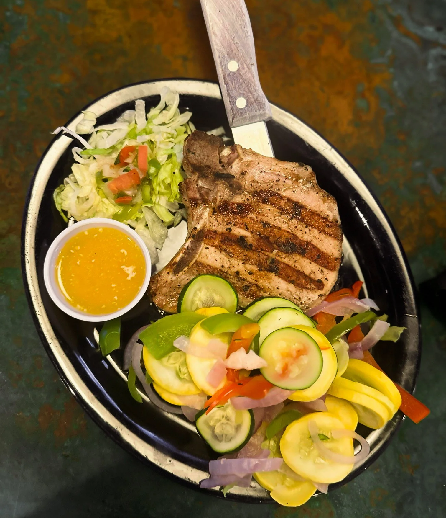 A plate with grilled pork chop, fresh sliced cucumbers, yellow zucchini, and a side of salad with shredded lettuce, tomatoes, and a small cup of yellow dressing.