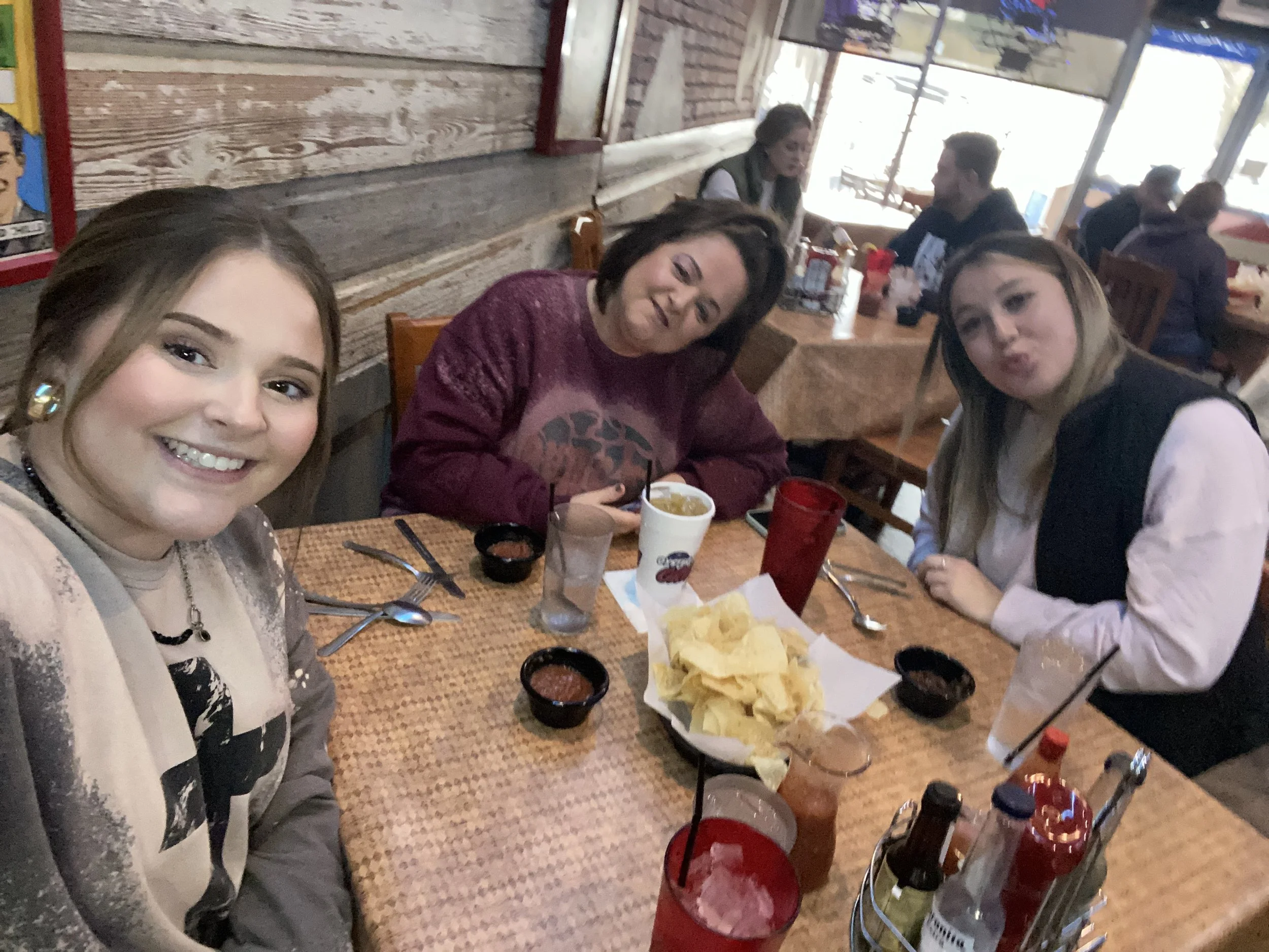 Three women sitting at a restaurant table with chips, salsa, drinks, and condiments, smiling for a photo.