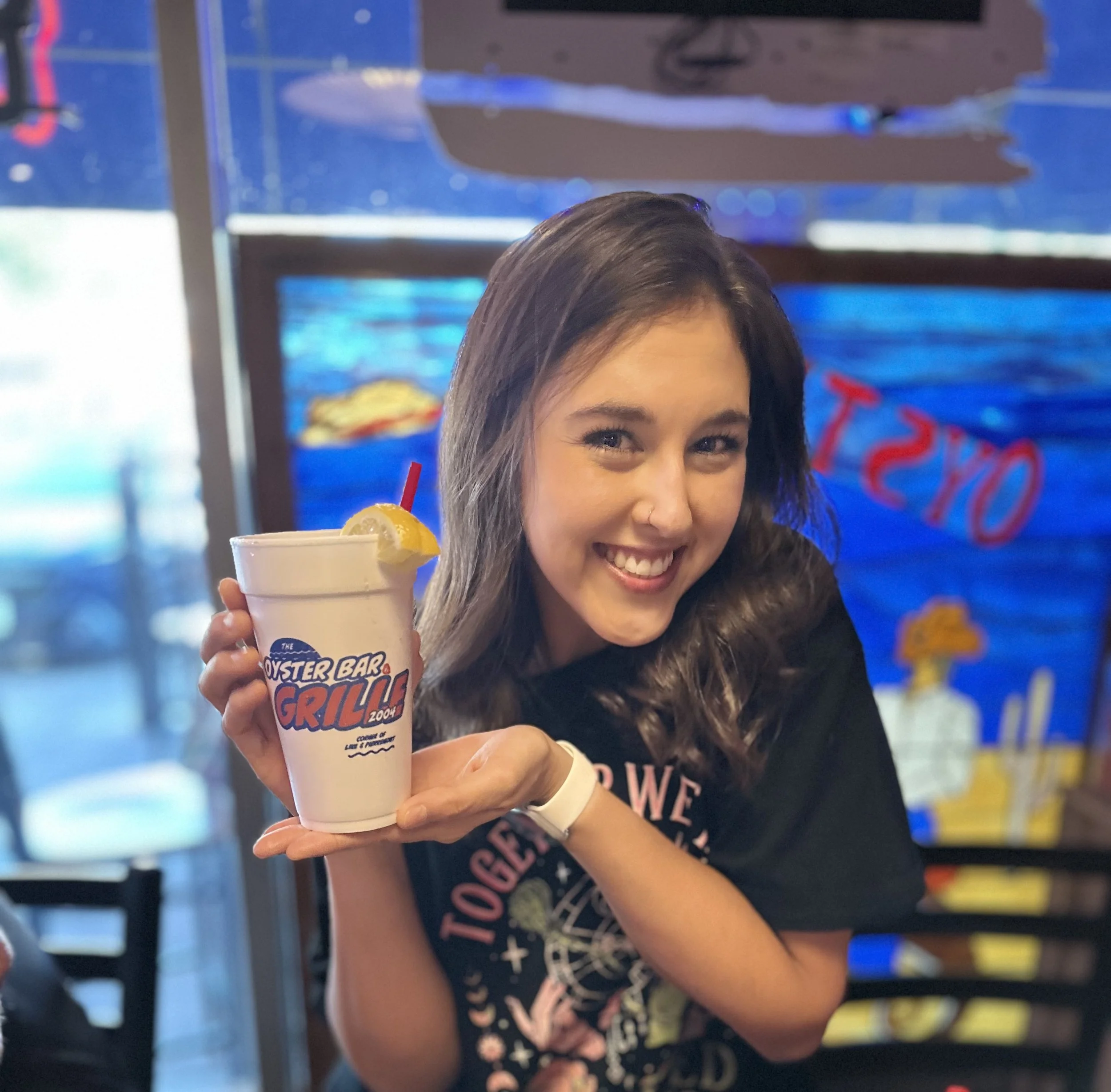 A woman with brown hair holding a white cup with a lemon slice and a cherry, inside a restaurant or cafe with blue, yellow, and red signs in the background.