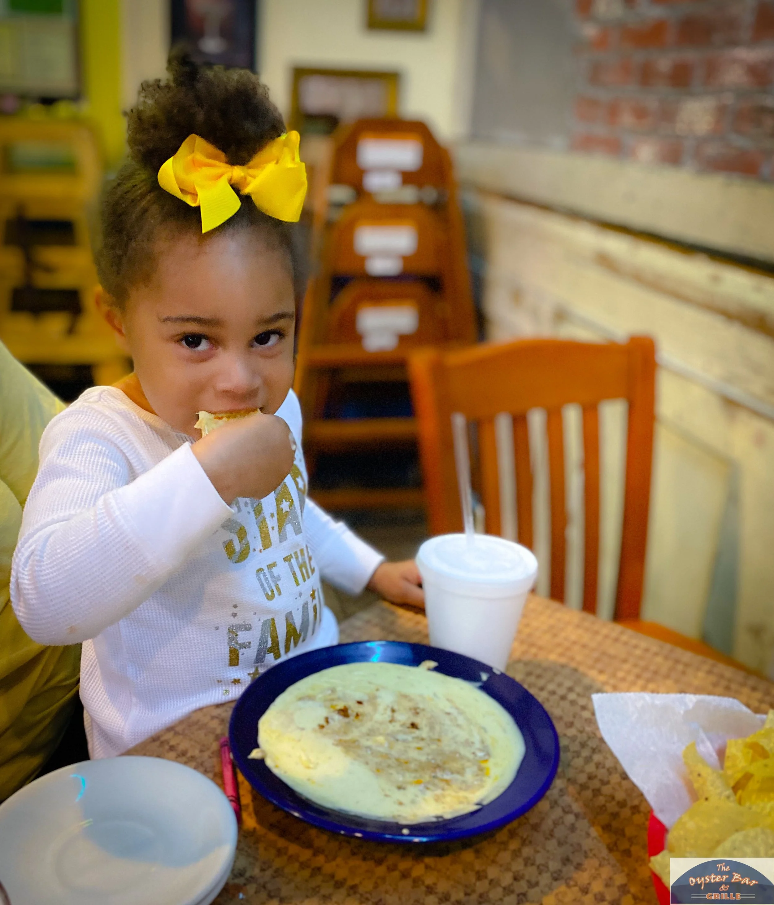 A young girl with curly hair tied up with a yellow bow, eating a slice of pizza at a restaurant. On the table in front of her are a large pizza, a white styrofoam cup, and a small empty white bowl. The background has yellow and brick wall elements an