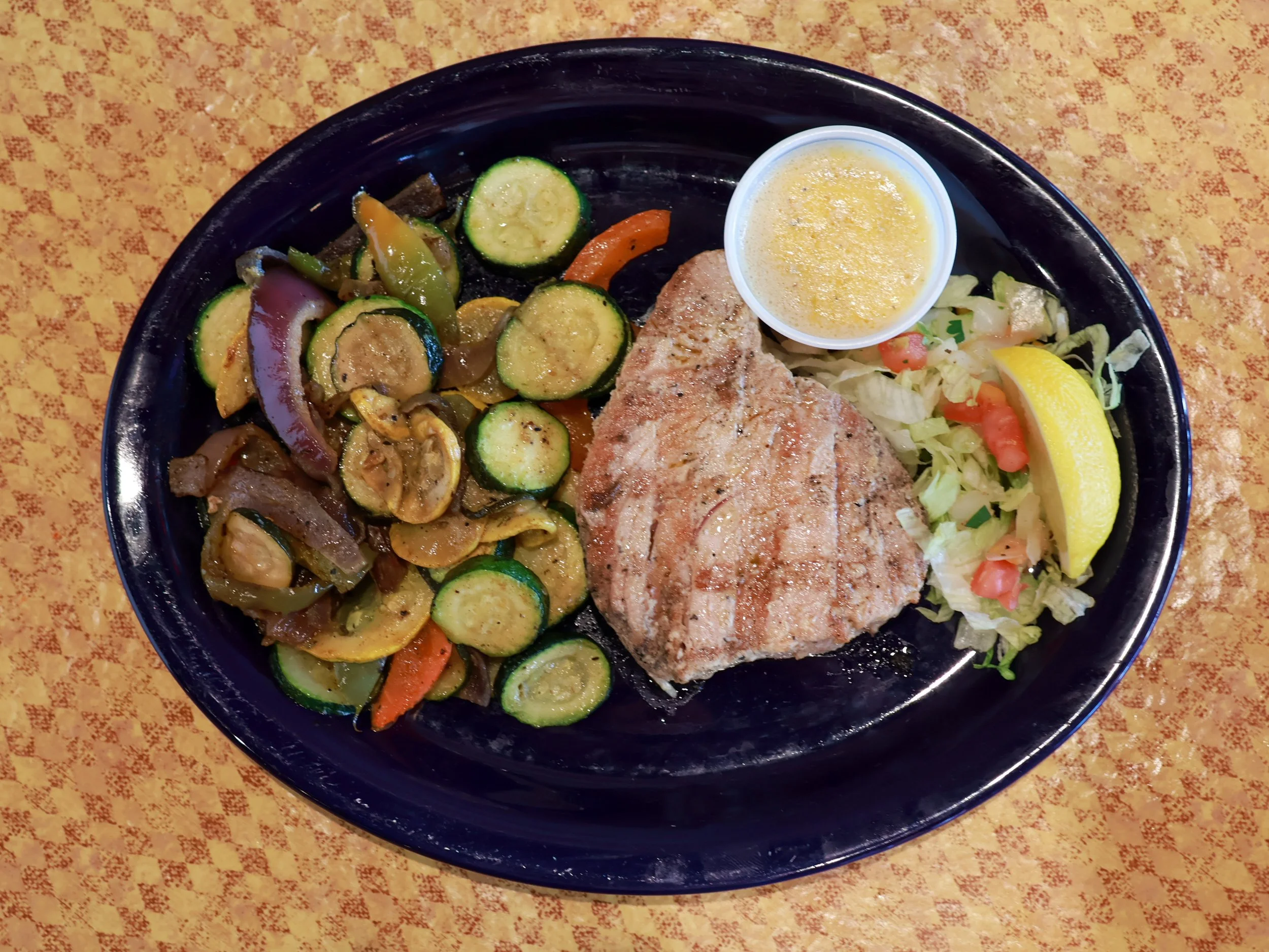 A plate of grilled fish with sautéed zucchini, bell peppers, onions, and a side salad with a lemon wedge and a small cup of melted butter.