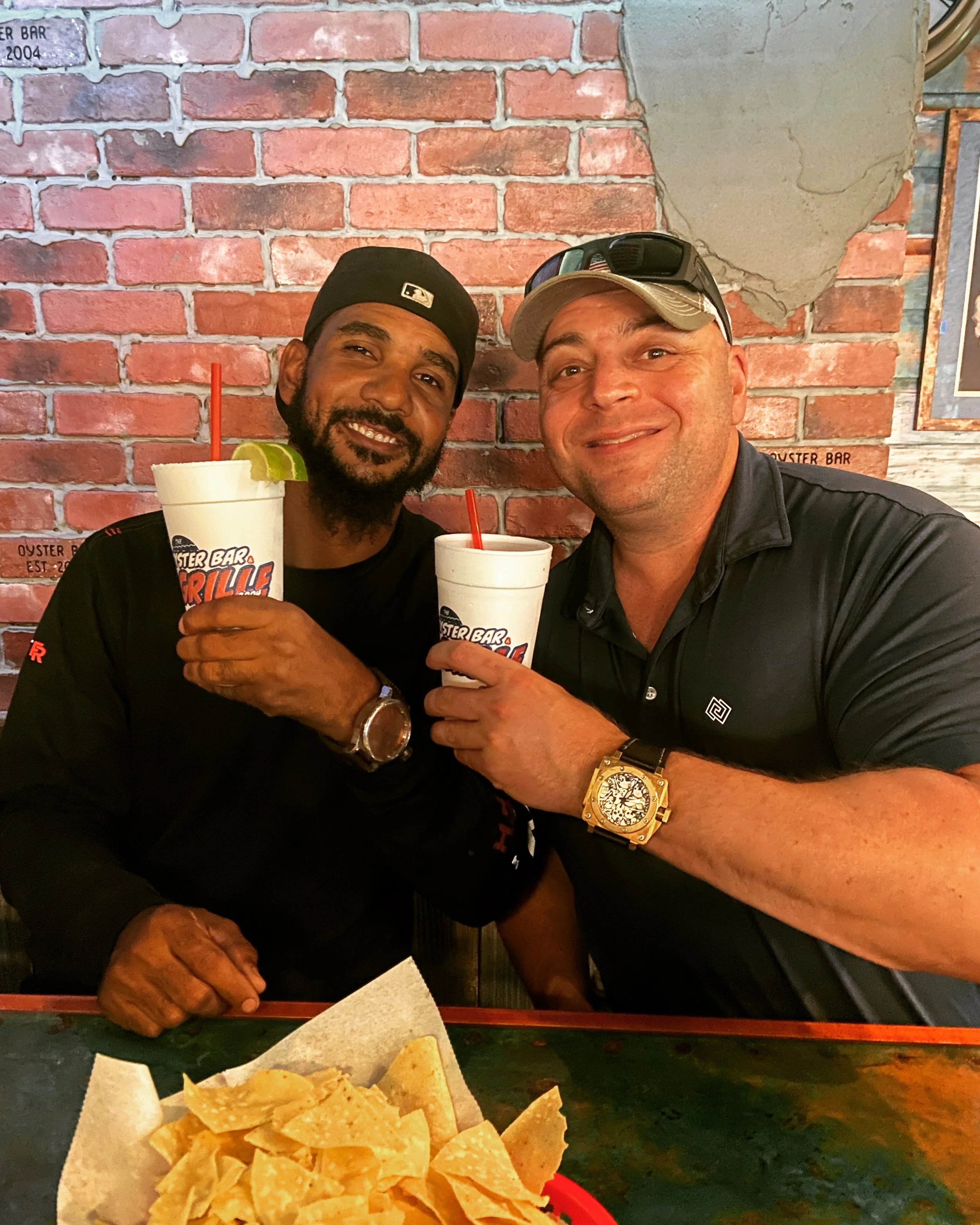 Two men smiling and holding drinks in a restaurant with a brick wall background. One man is wearing a black cap and the other a beige cap with sunglasses on top. They are sitting at a table with a basket of tortilla chips in the foreground.