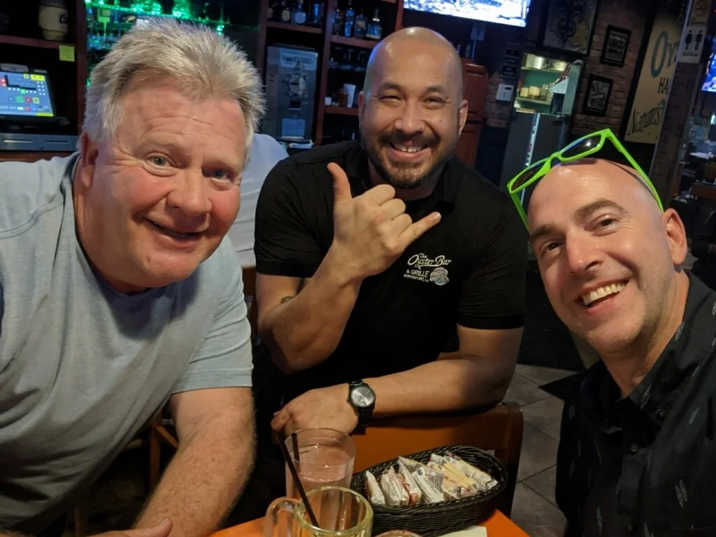Three men smiling and sitting at a table in a bar or restaurant. The man on the left has light hair, the man in the middle has a bald head and a goatee, and the man on the right has a shaved head with bright green sunglasses resting on his head. Ther