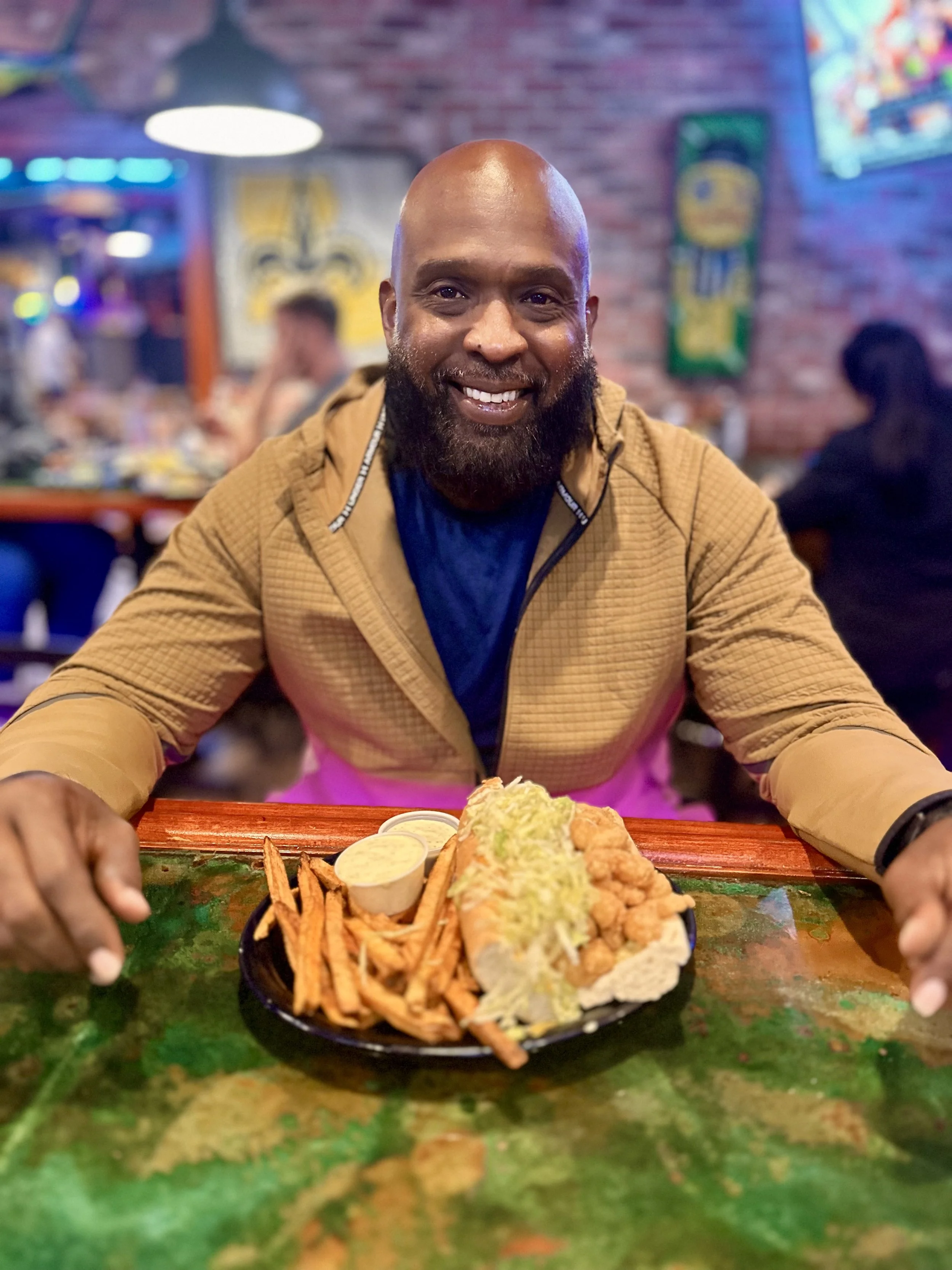 A smiling man with a beard seated at a restaurant table with a plate of food, including fries, shredded lettuce, and dipping sauces, in front of him. The background shows a brick wall, neon lights, and colorful decorations.