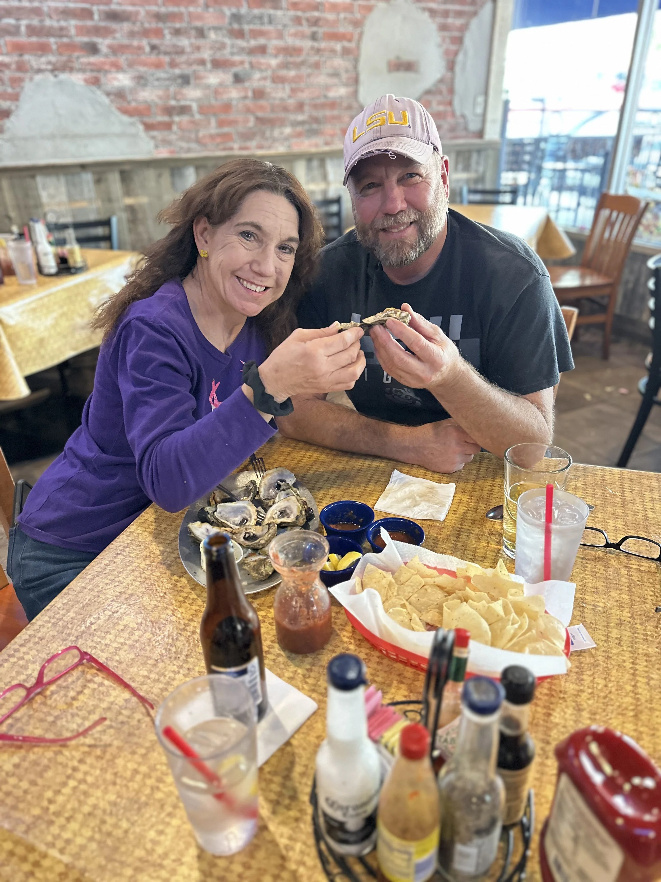 A man and a woman smiling and enjoying oysters at a restaurant table, with various condiments, drinks, and a basket of tortilla chips on the table.