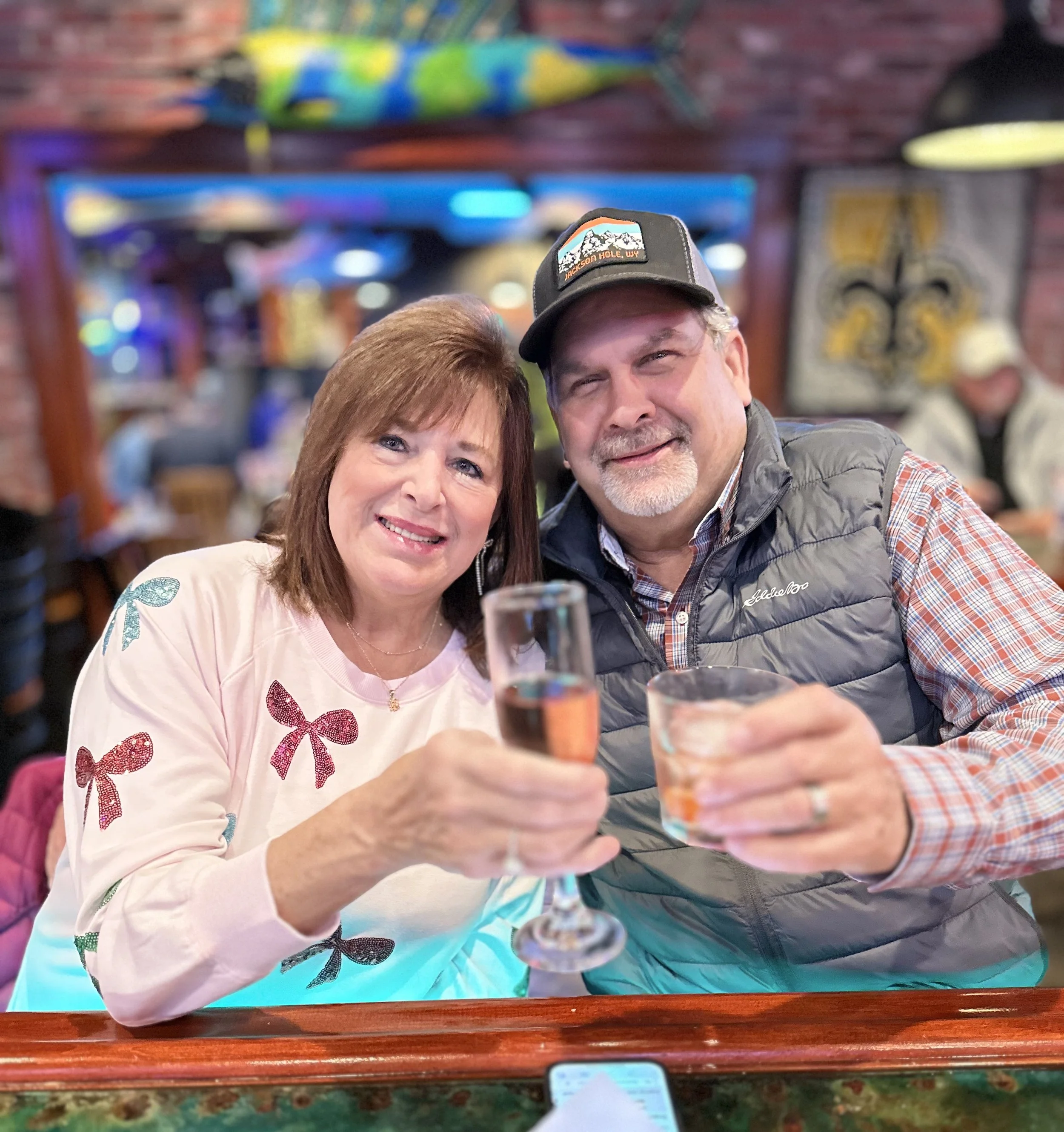 Two people, a woman with red hair and a man with gray hair, sitting in a colorful restaurant. They are smiling and taking a selfie. The woman is wearing a pink sweater with butterfly designs and the man is wearing a gray jacket and a baseball cap wit