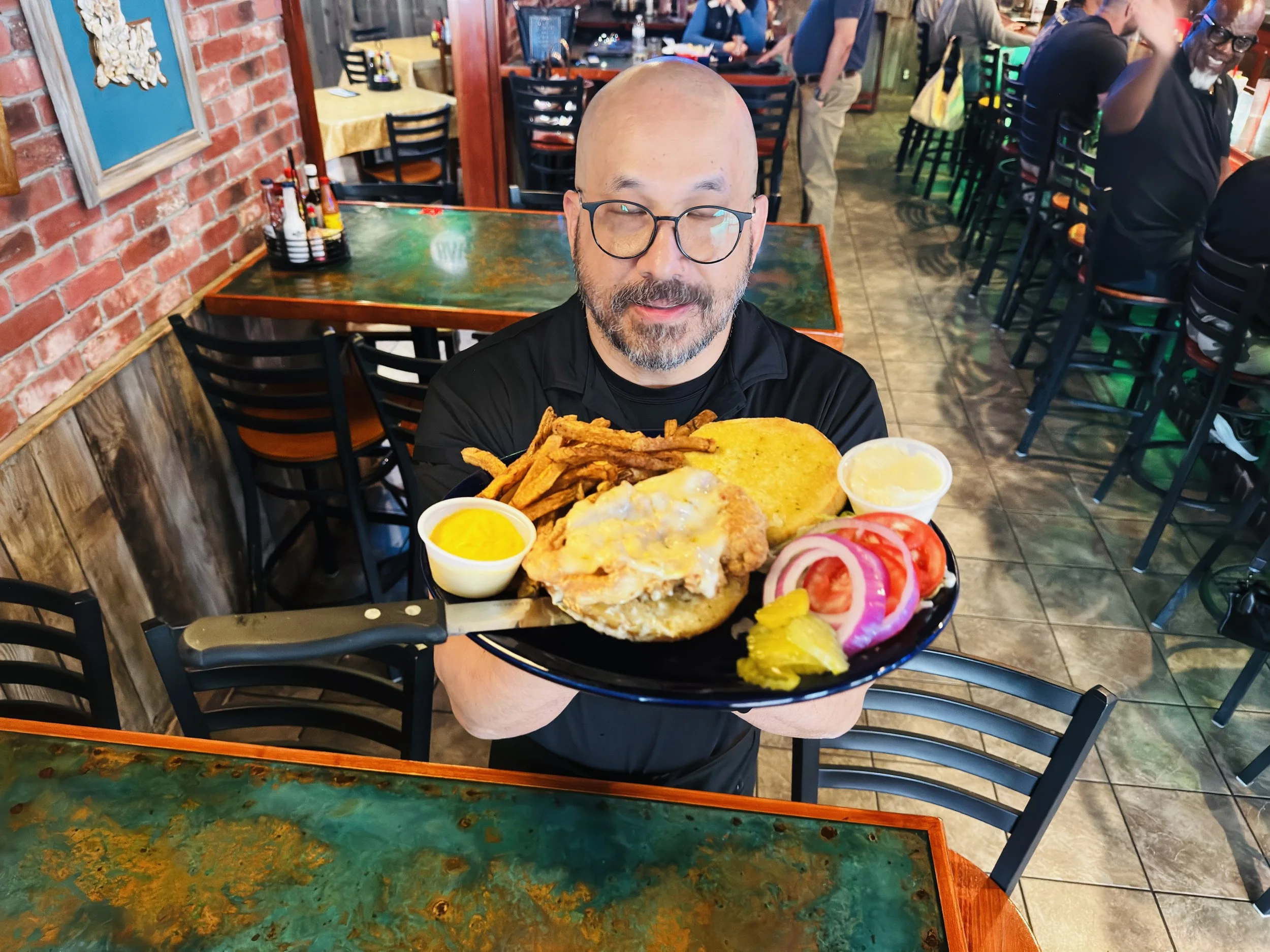A man with glasses and a beard holding a large plate of fried chicken sandwich, Hand cut fries, tomato slices, pickles, and two cups of dipping sauces inside a restaurant.