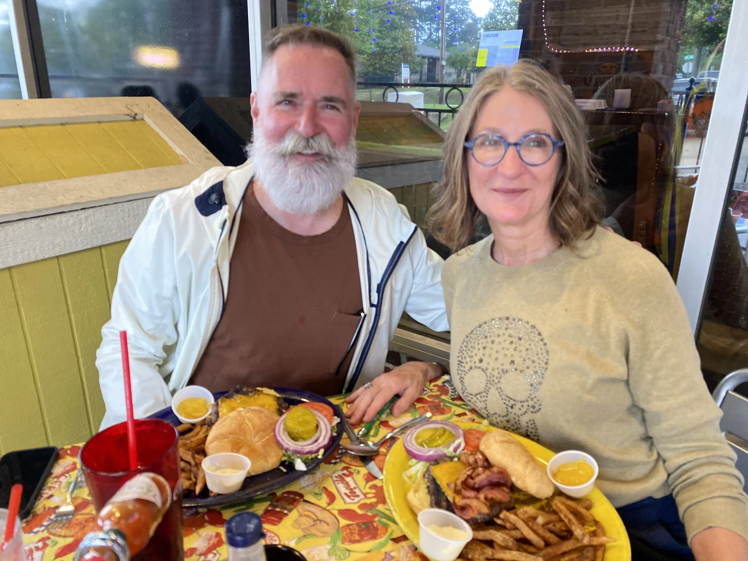 A man with a beard and a woman with glasses sitting at a table with a large plate of Mexican food, including tacos, pickles, onions, and sides. They are in a restaurant with windows showing a street outside.