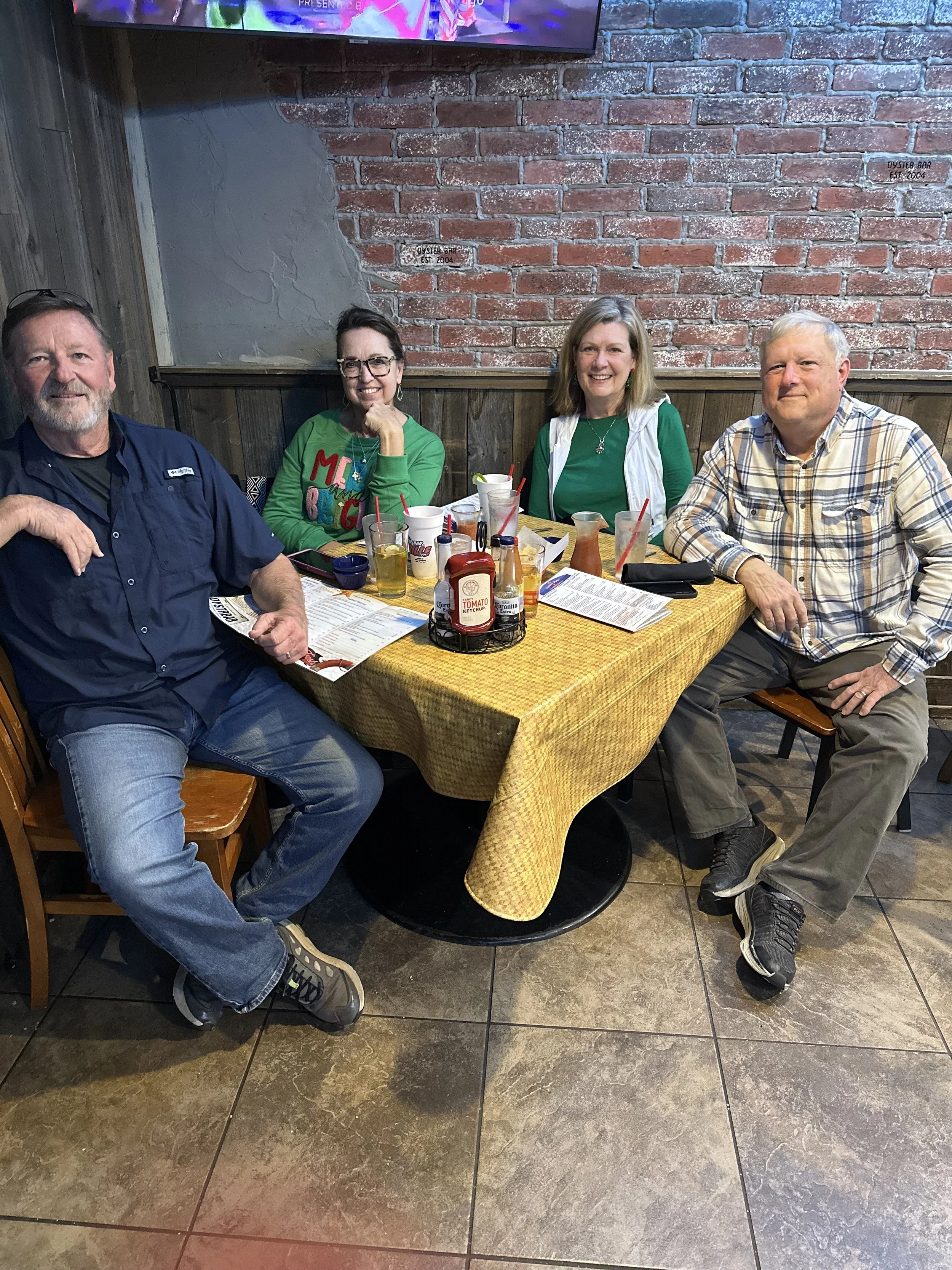 Four adults sitting at a table in a restaurant or bar with a brick wall background. They are smiling and enjoying drinks, with condiments and menus on the table.