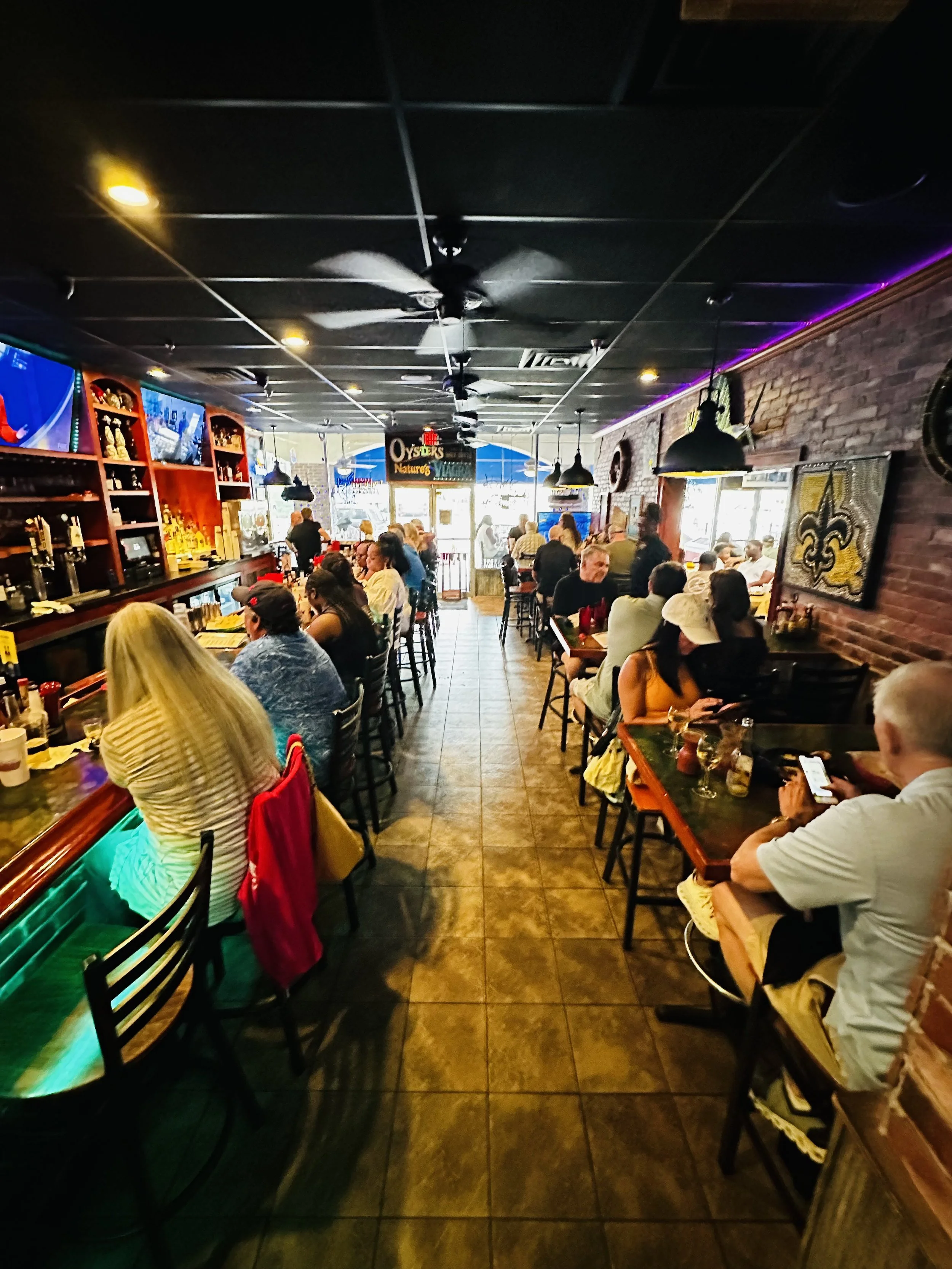 Inside a busy restaurant with crowded tables, patrons, brick walls, television screens, and a newly remodeled bar area. Local artists display artwork on walls.