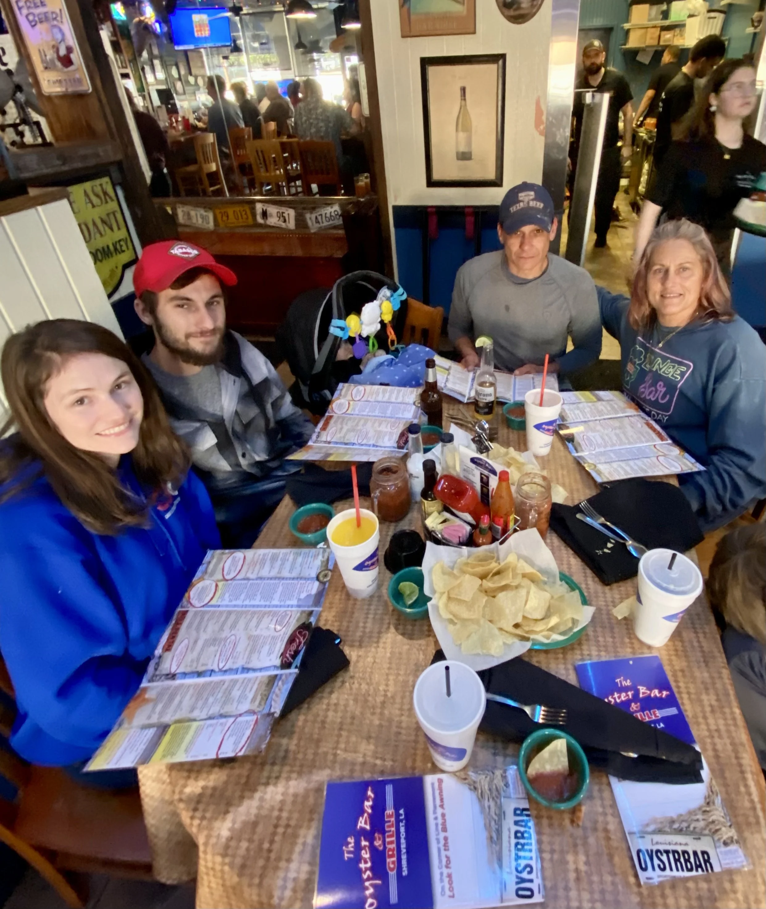 A group of people sitting around a restaurant table with food, drinks, and menus; bar and restaurant interior in the background.