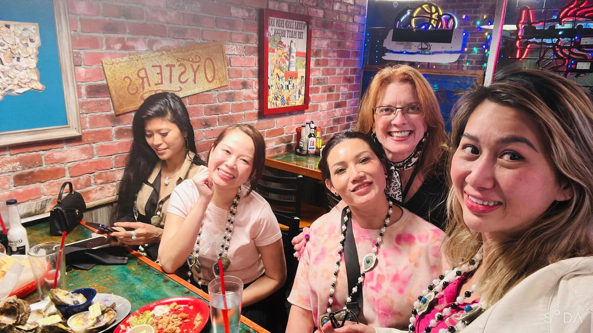 A group of five women smiling at a restaurant or bar with a brick wall background and colorful string lights, with some dishes and drinks on the table in front of them.