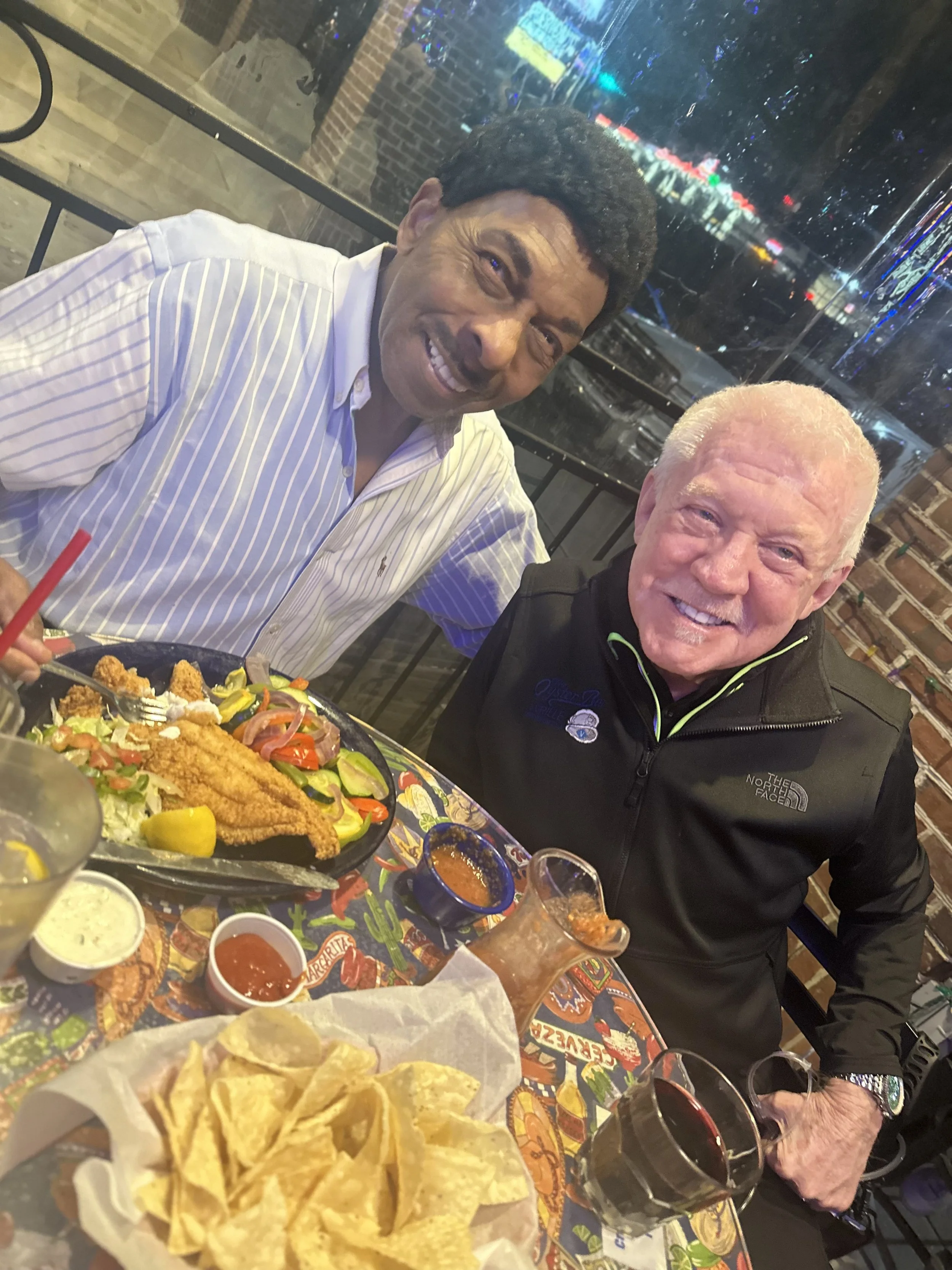 Two men smiling at a restaurant table with Mexican food, including nachos and fried chicken, and drinks. The background shows a window with city lights.