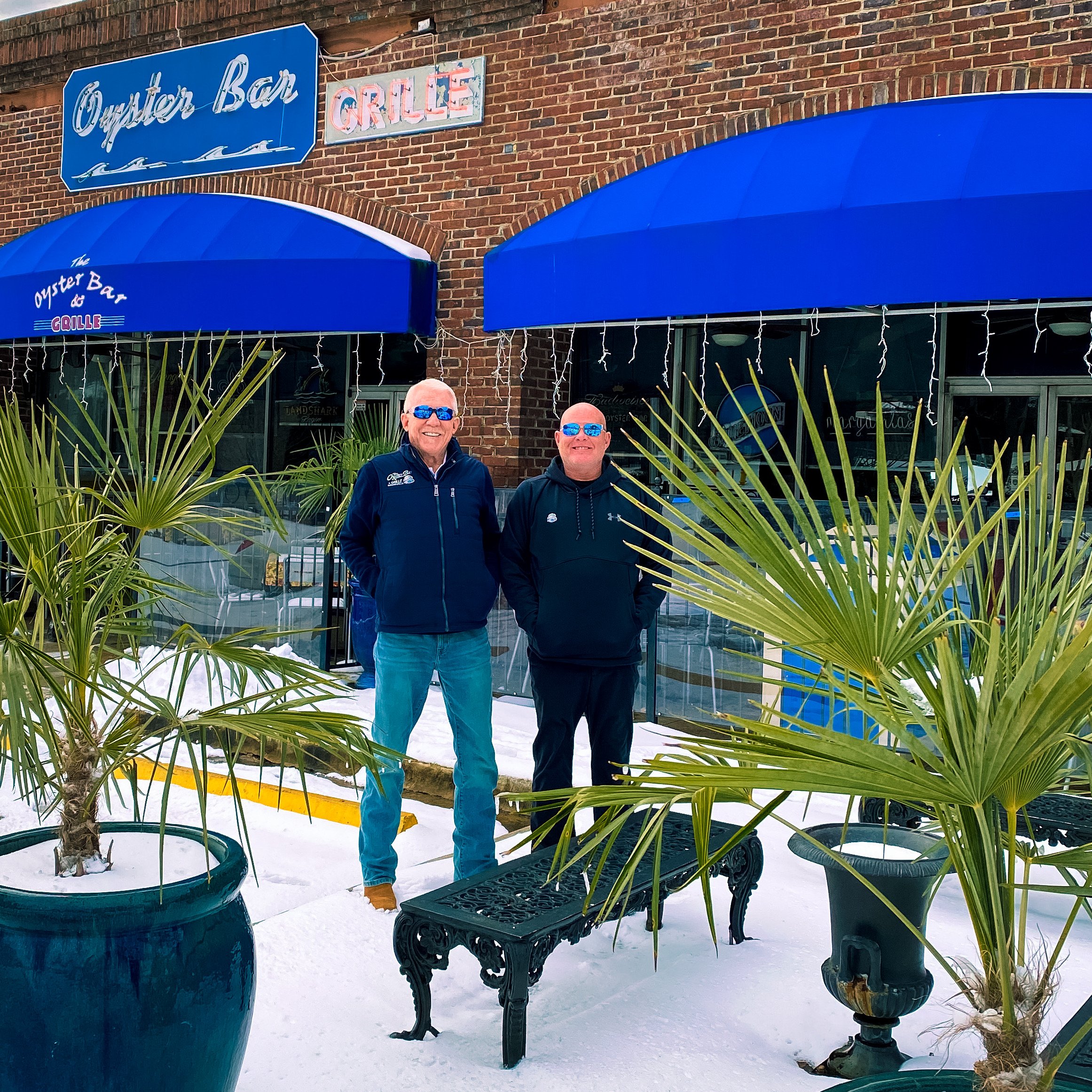 Two men are standing outside in front of potted palm trees on a snowy day. Behind them is a brick building with blue awnings and signs reading 'Oyster Bar' and 'Grille'