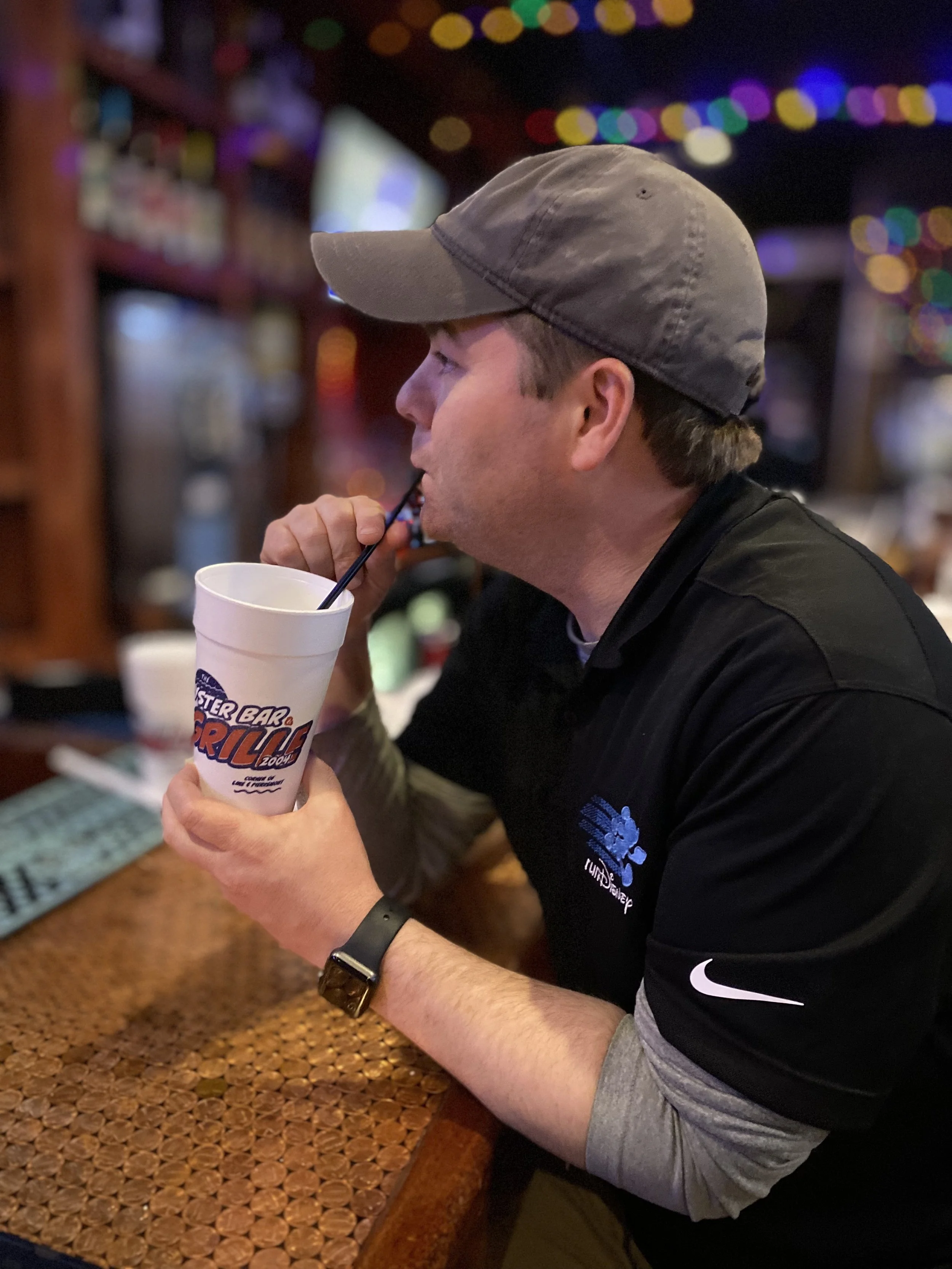 A man wearing a gray baseball cap and a black shirt with a blue and white logo sits at a bar counter, drinking from a Styrofoam cup with a straw. The background features colorful blurred lights.