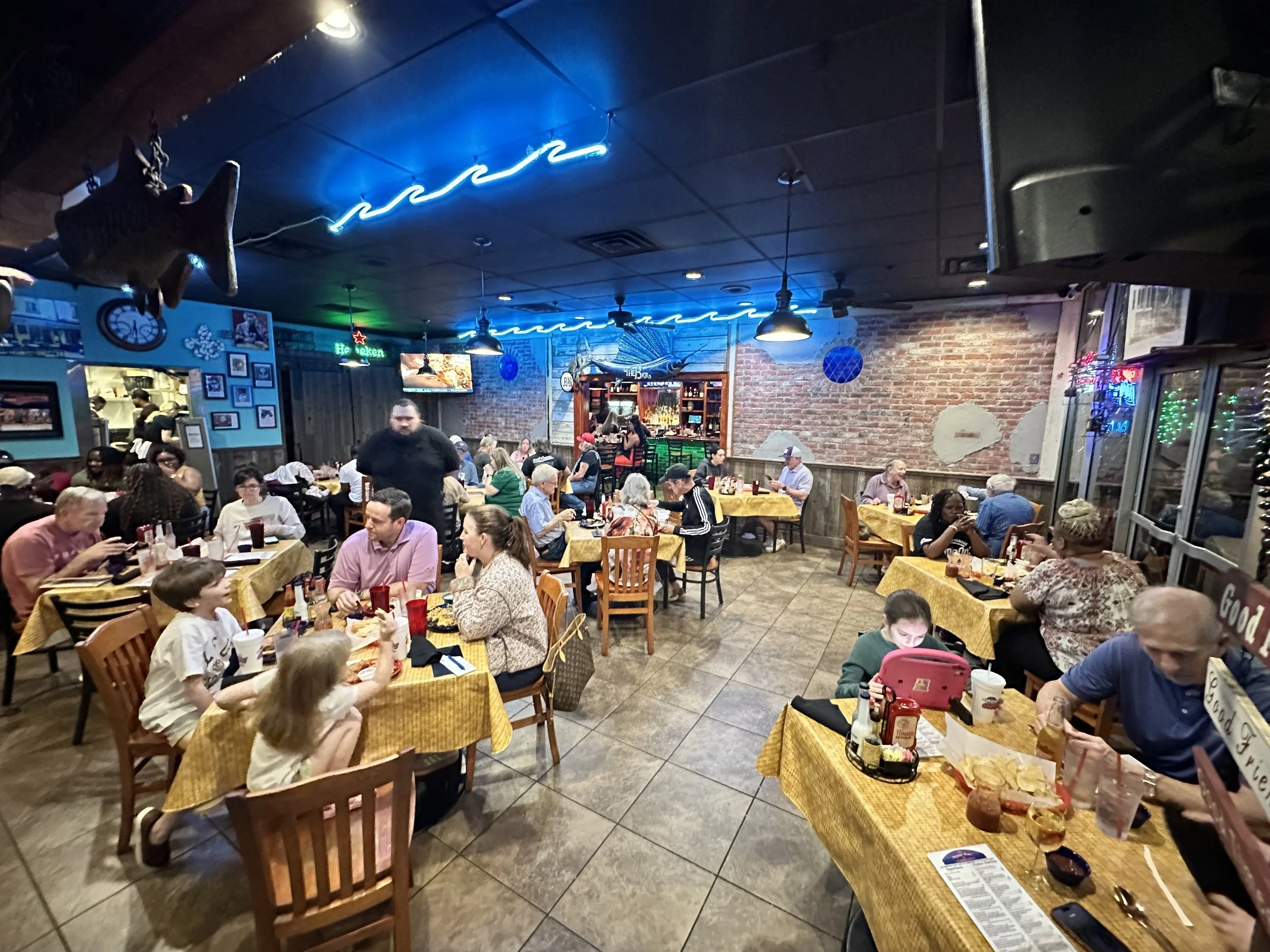 Interior of a lively restaurant with many diners at tables covered with yellow checkered tablecloths, some eating, some using phones, and a bar area in the background. Neon lights and brick walls add to the cozy ambiance.