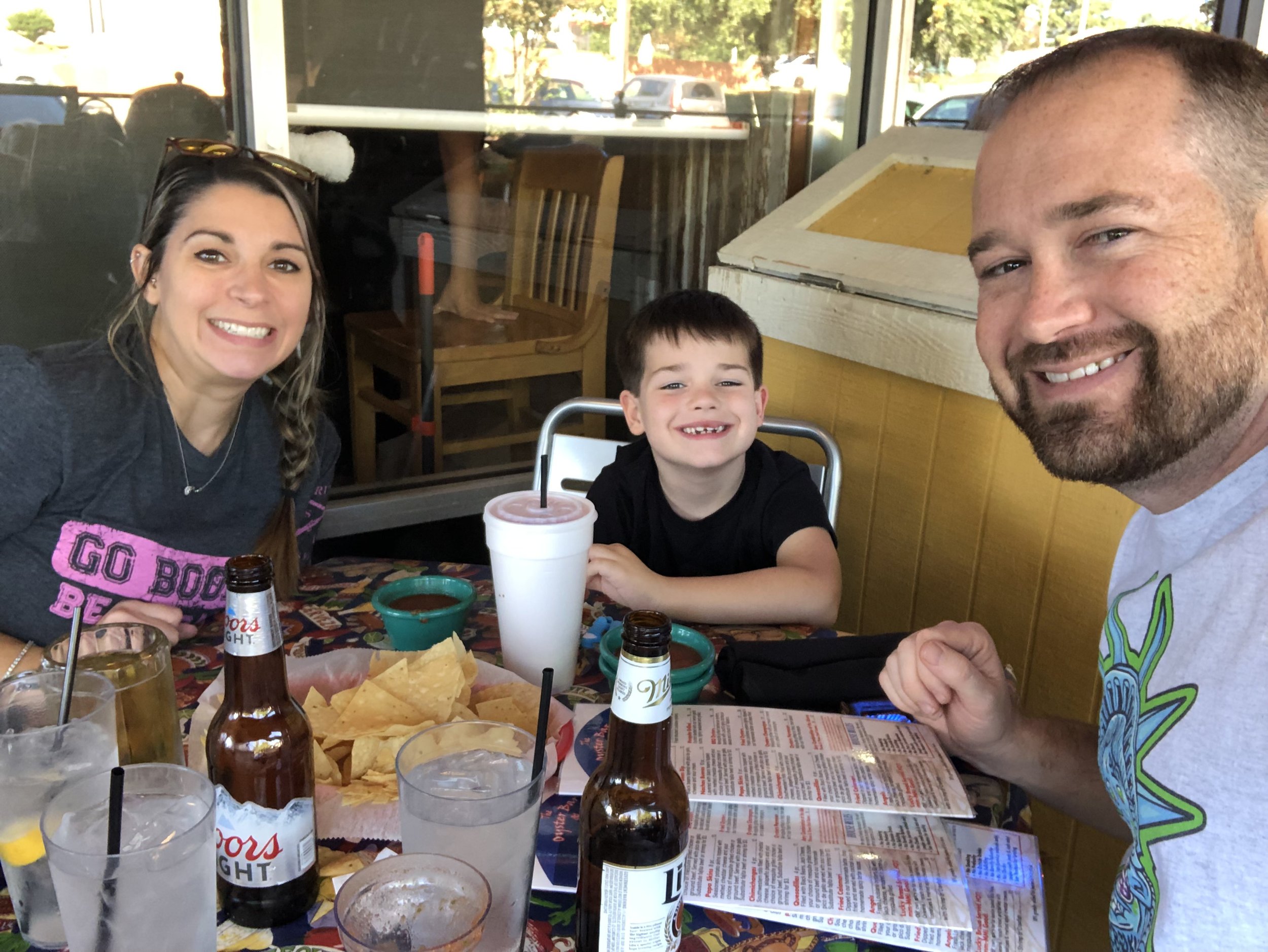 Family of three sitting at a restaurant table with drinks, chips, and salsa, smiling at the camera.