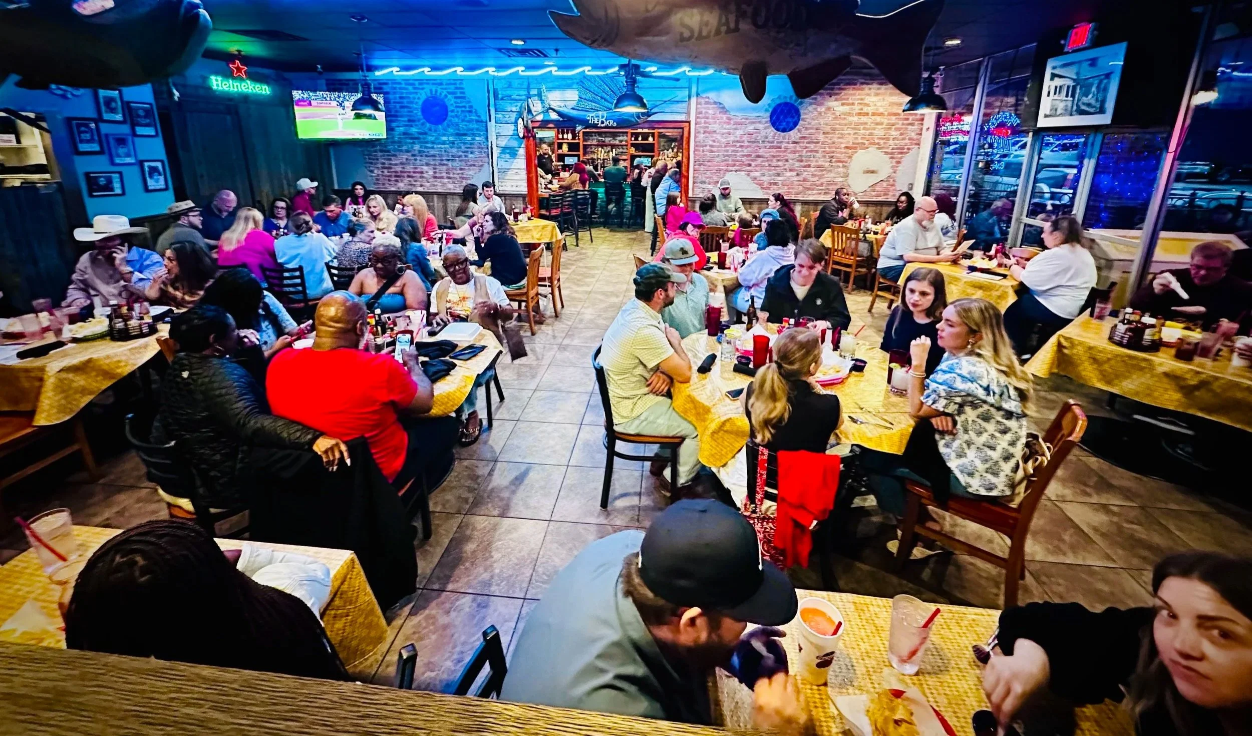 A lively restaurant filled with diverse patrons sitting at tables with yellow tablecloths, enjoying meals and conversations. The decorated brick walls feature neon signs and framed pictures, with a bar area in the back. The atmosphere is vibrant and casual.