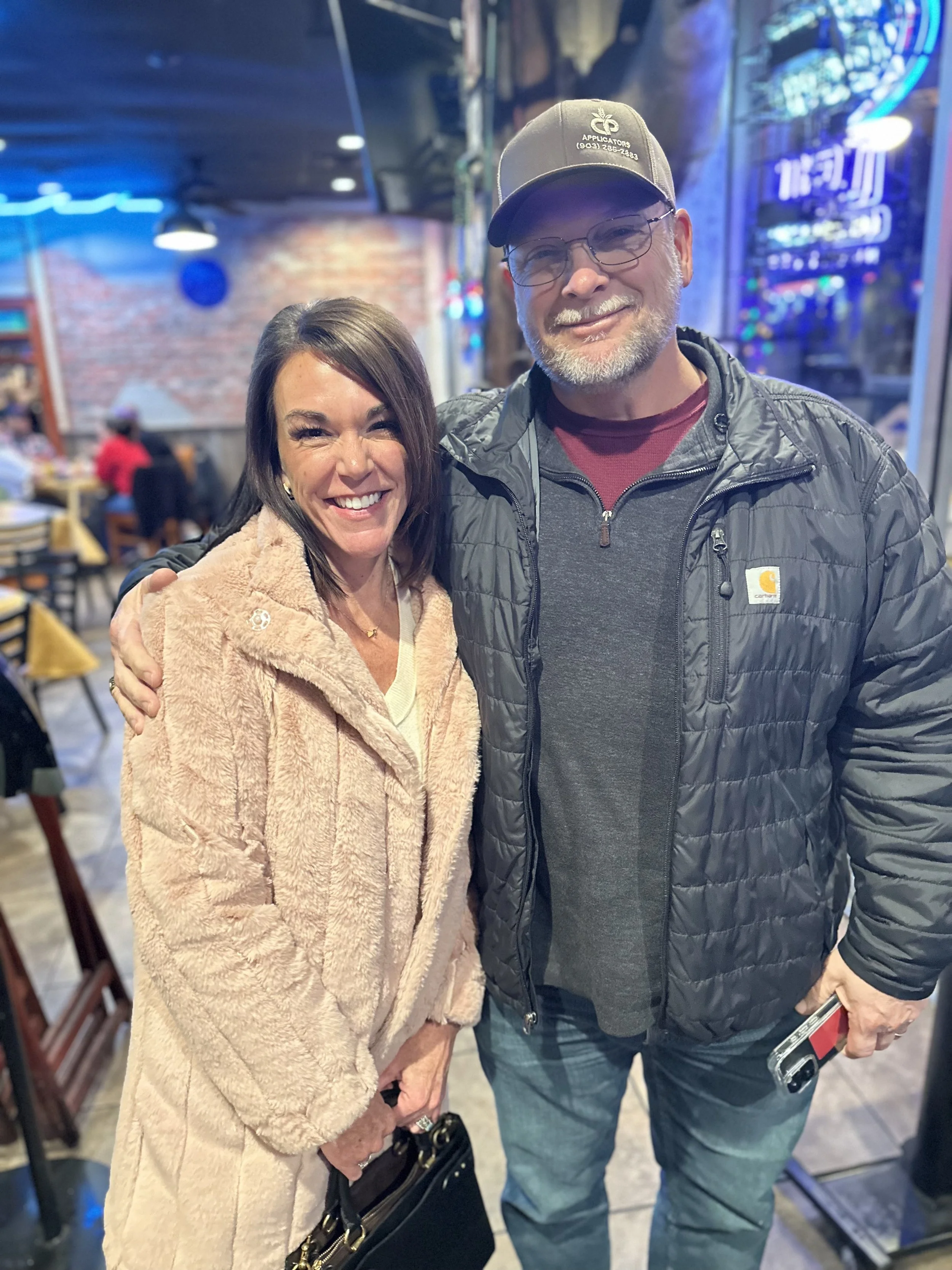 Two people, a woman and a man, smiling and posing together in a restaurant. The woman has dark hair and is wearing a beige fuzzy coat, holding a black purse. The man has glasses, a gray beard, and is wearing a gray cap, a gray jacket, and a maroon sh