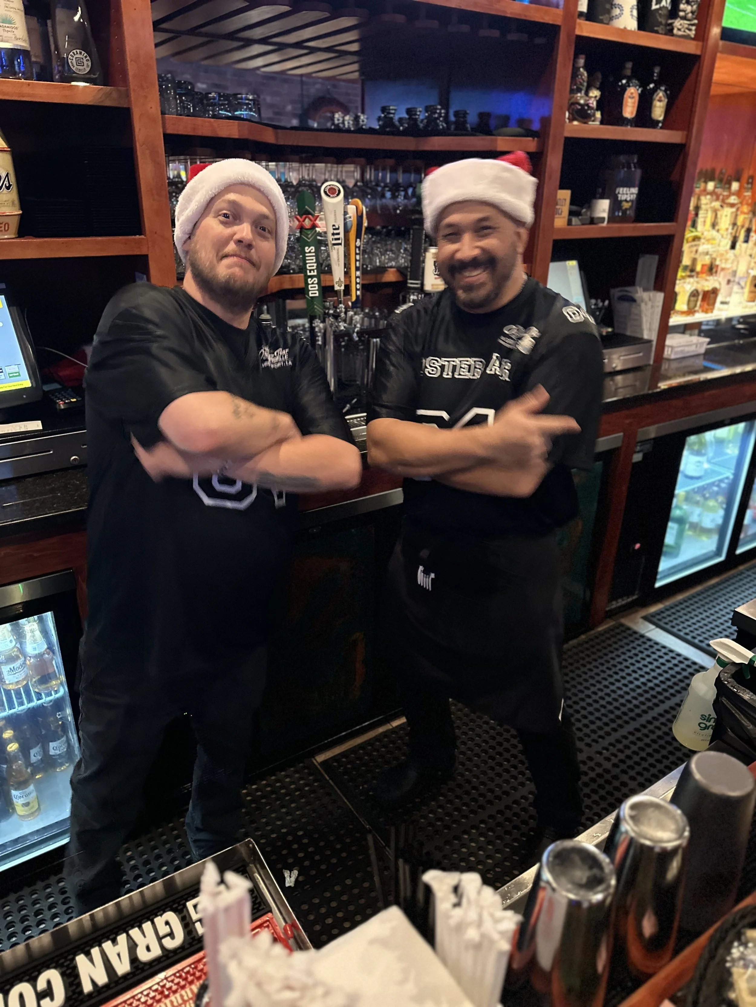 Two bartenders wearing Santa hats behind a bar with alcohol bottles and taps, smiling and posing with crossed arms.
