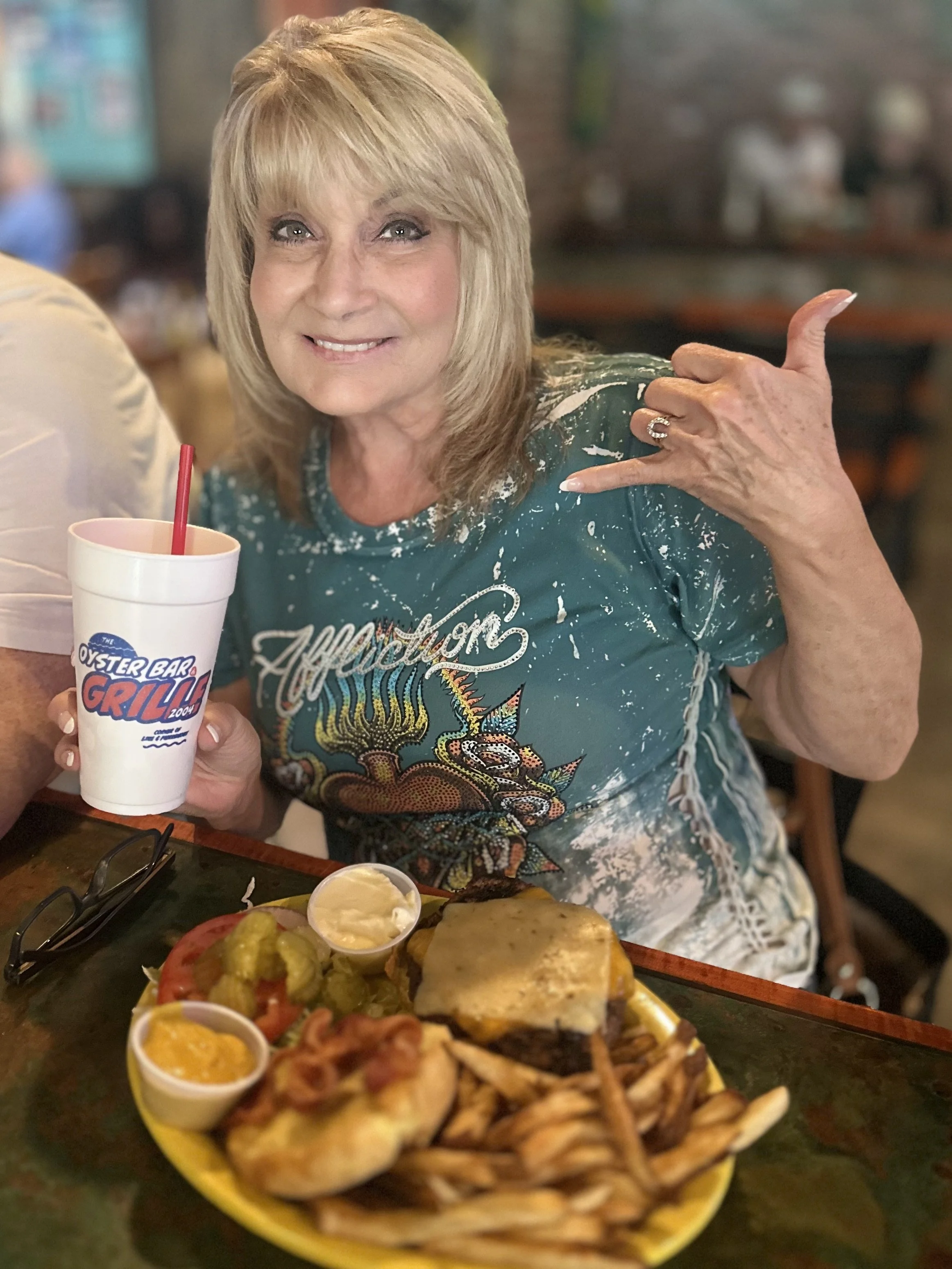 A woman with blonde hair smiling and making a hand gesture at a restaurant table with food, including a pizza, dips, and a large drink.