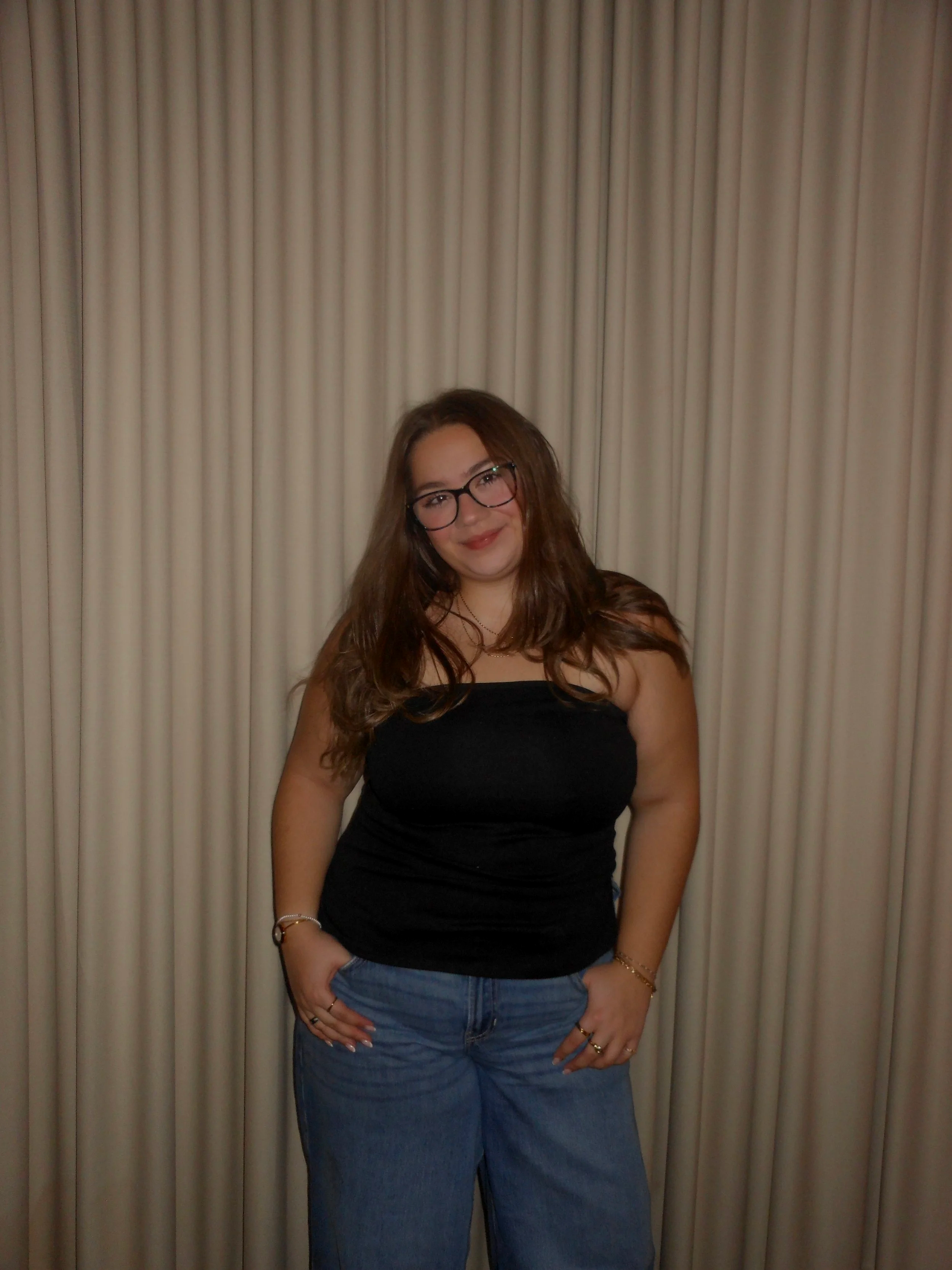 A young woman with long brown hair, glasses, and light skin, standing in front of a beige curtain. She is wearing a black strapless top, light blue jeans, and accessories including rings and bracelets. She is smiling and resting her hands on her hips.