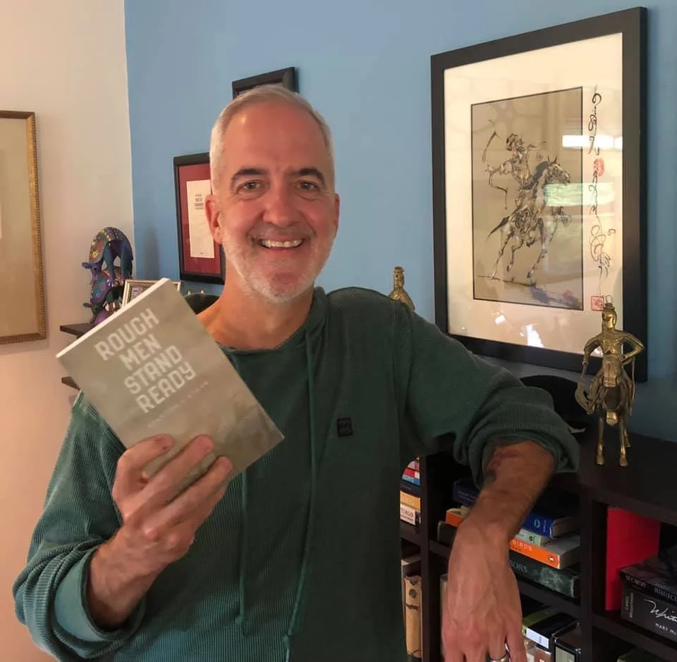 A smiling middle-aged man with gray hair and a beard holding a book titled "Rough Men Stand Ready" in a room with artwork and decorative items on shelves.