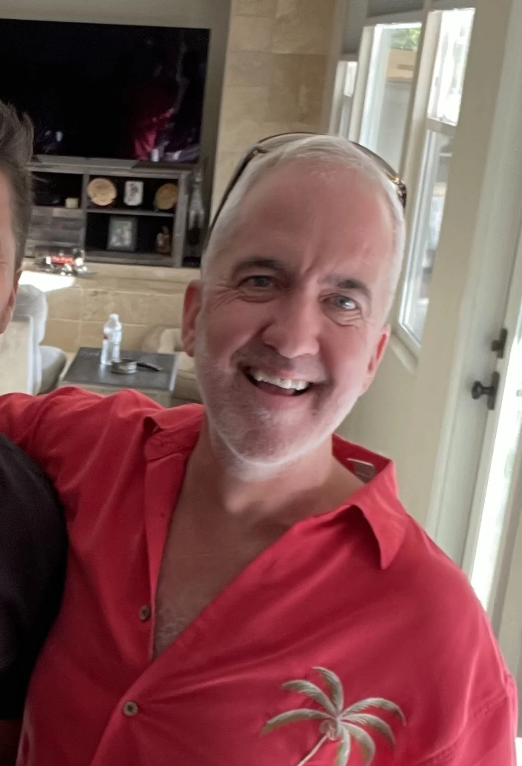 A smiling man with short, gray hair, wearing a red shirt with a palm tree embroidery, standing inside a house with beige walls and large windows.