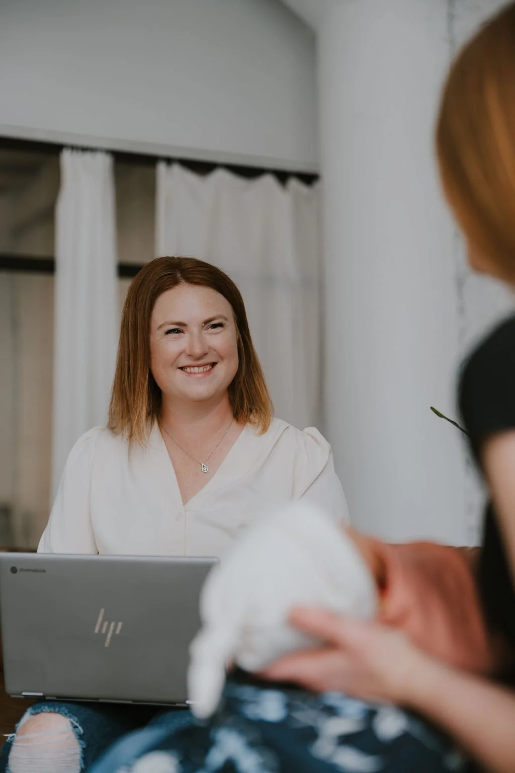 A woman with red hair smiling at another person holding a baby, in a room with white walls and curtains, sitting with a laptop in front of her.