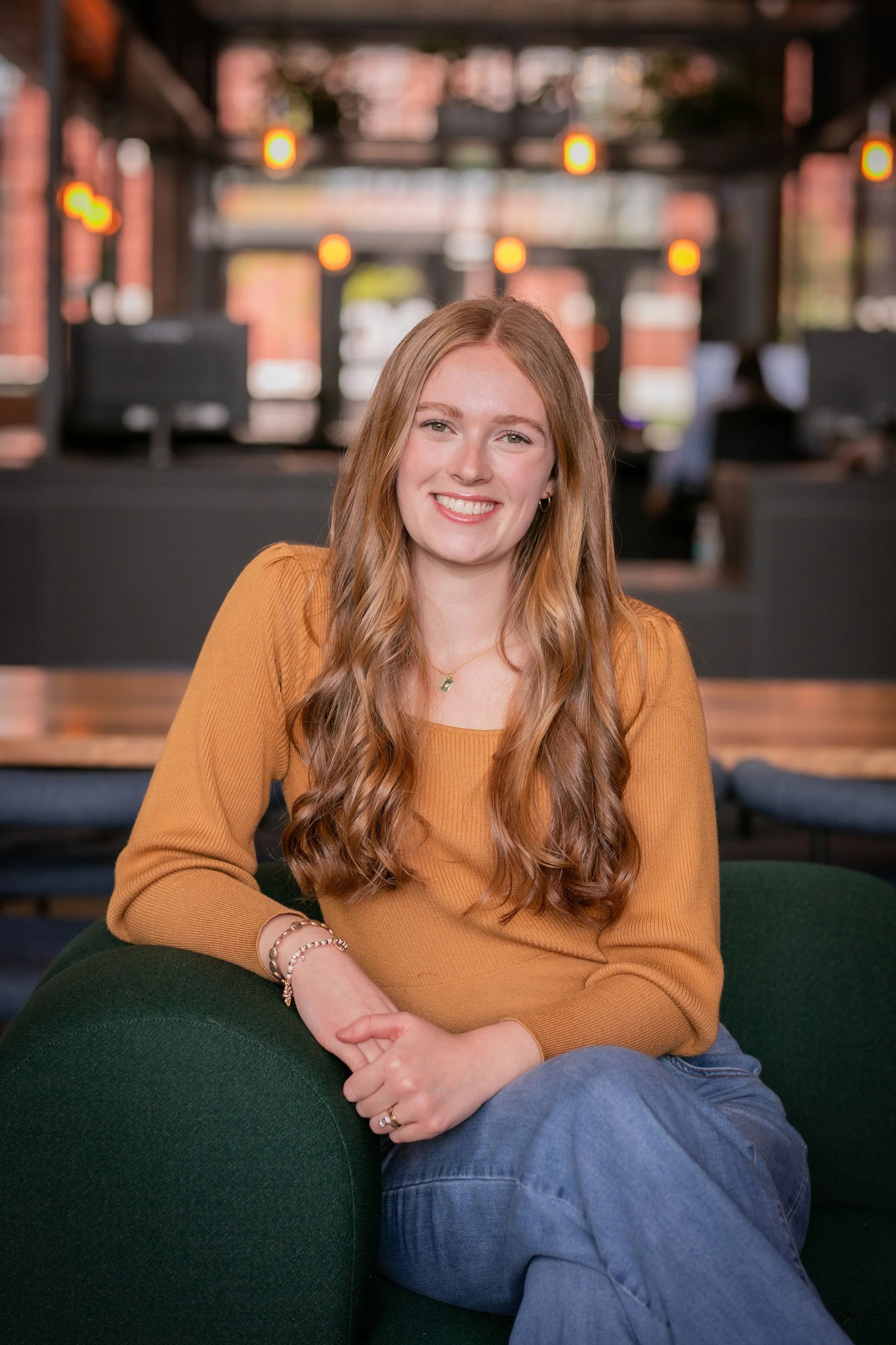 A young woman with long wavy red hair, wearing a mustard yellow sweater, sitting on a dark green couch with her hands clasped in her lap, smiling at the camera. The background shows a blurred indoor space with brick walls, open windows, and warm lighting.