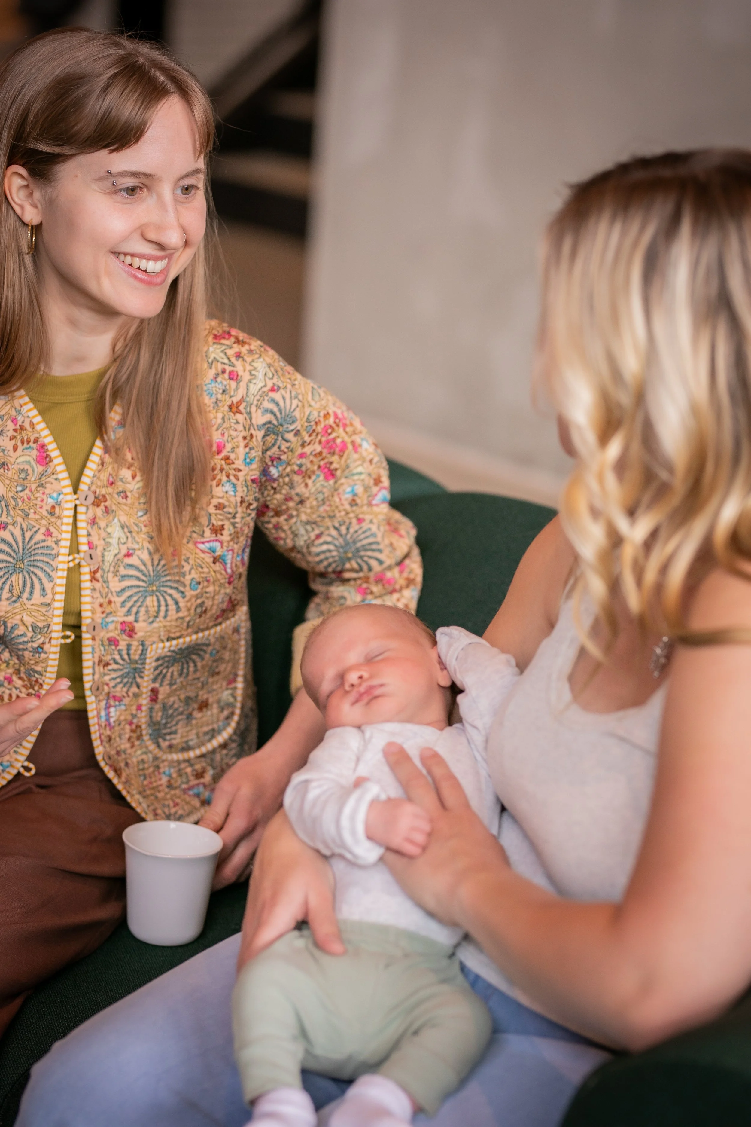 A woman with long blonde hair holding a sleeping baby while sitting on a couch, with a woman with long brown hair and a patterned jacket smiling and sitting nearby.