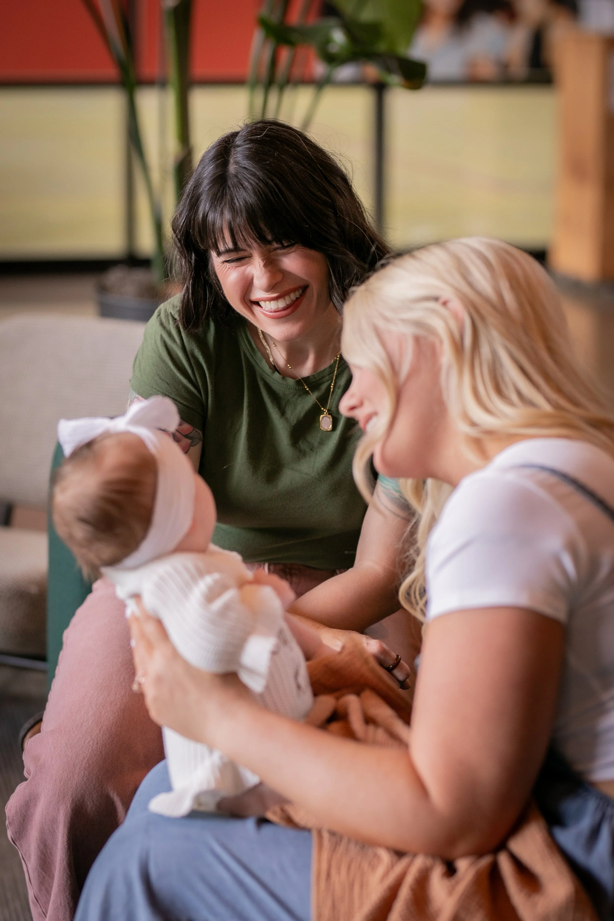 Two women and a baby, one woman with dark hair laughing and the other with blonde hair. The baby, wearing a white headband, is being held and looks at the woman with dark hair. The women appear to be enjoying a happy postpartum.