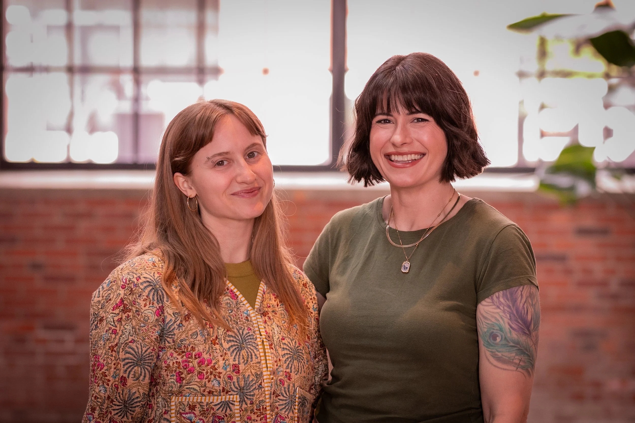 Two female postpartum doulas standing close, smiling at the camera in an indoor setting with large windows and brick wall background.
