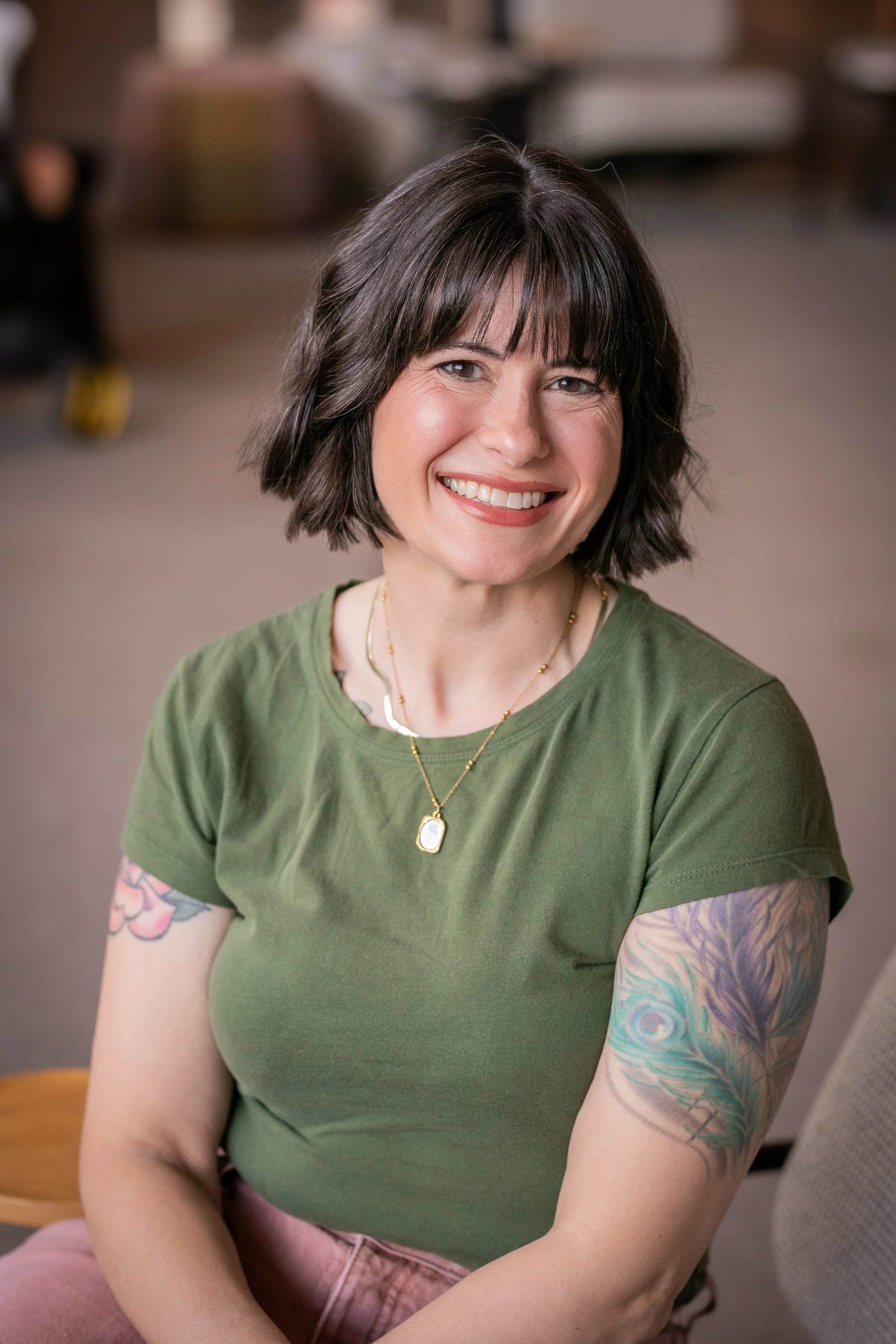A woman with dark hair and bangs, wearing a green t-shirt and gold jewelry, smiling at the camera. She has tattoos on her arm, and the background appears to be an indoor setting with blurred furniture.