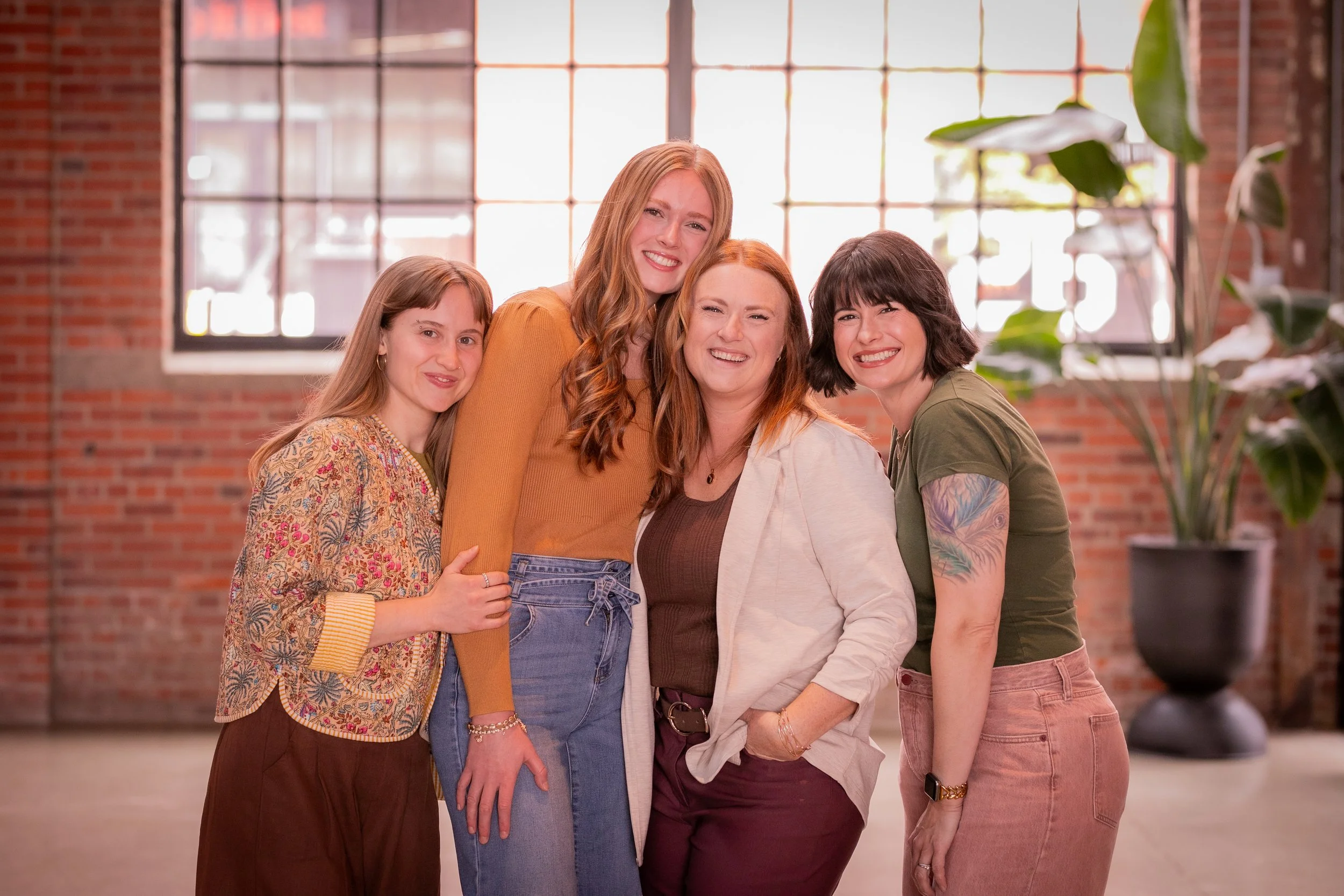 Group of five women smiling and posing together indoors with large windows and a brick wall in the background.