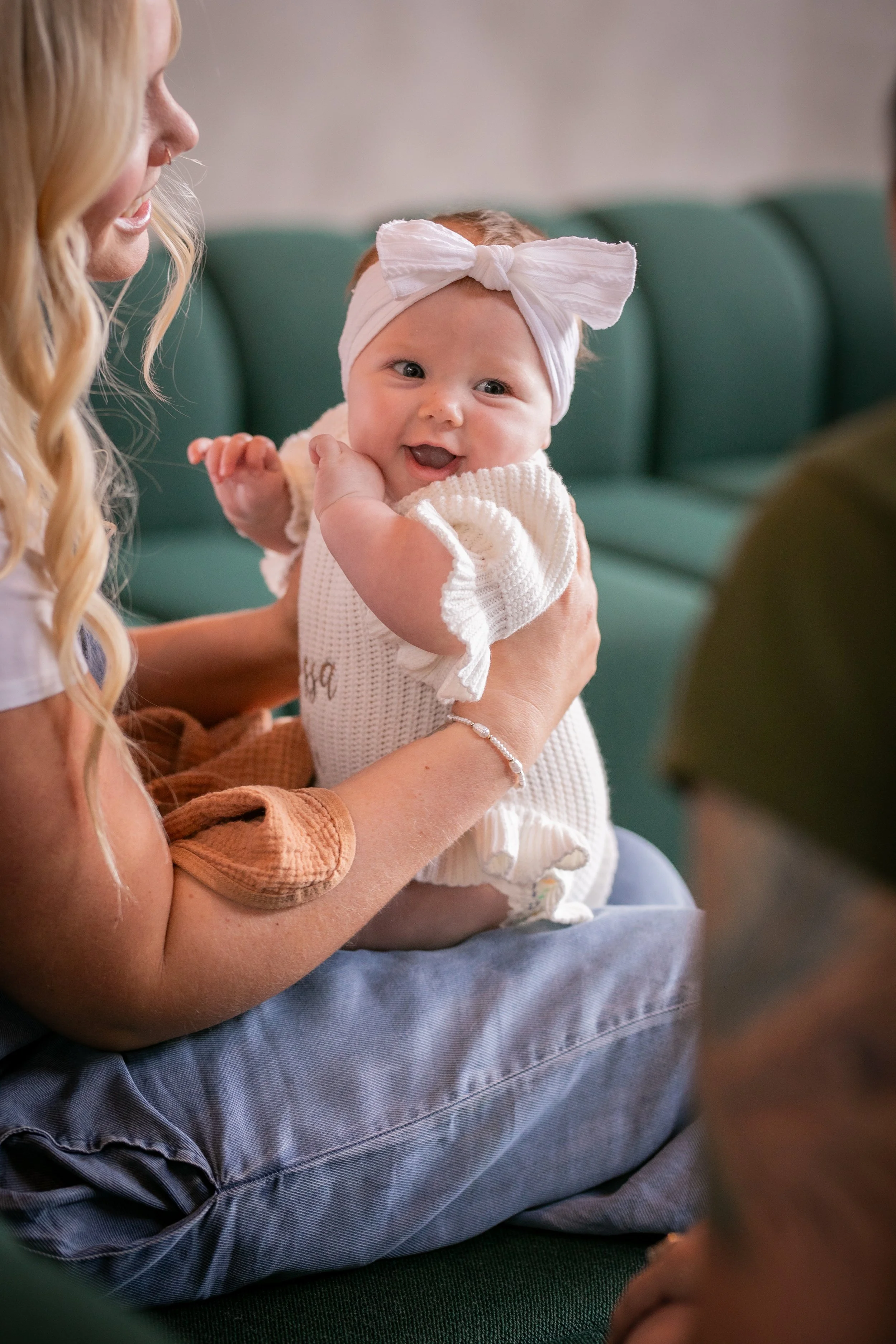 A woman holding a smiling baby girl with a white headband and cream-colored sweater.
