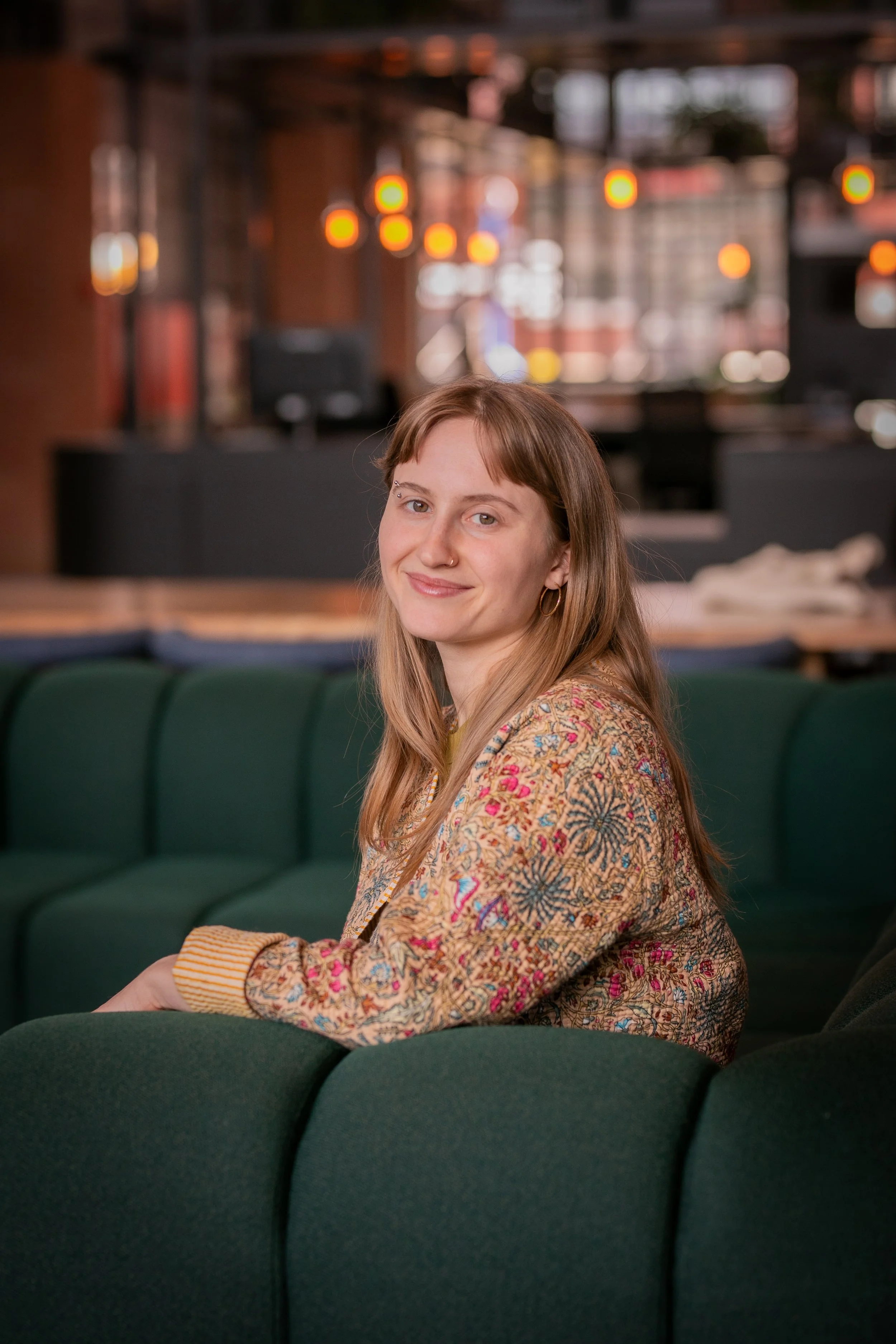 A woman with long light brown hair, wearing a patterned jacket and a yellow shirt, sitting on a green couch in a cozy, dimly lit space with warm lights and wooden decor.