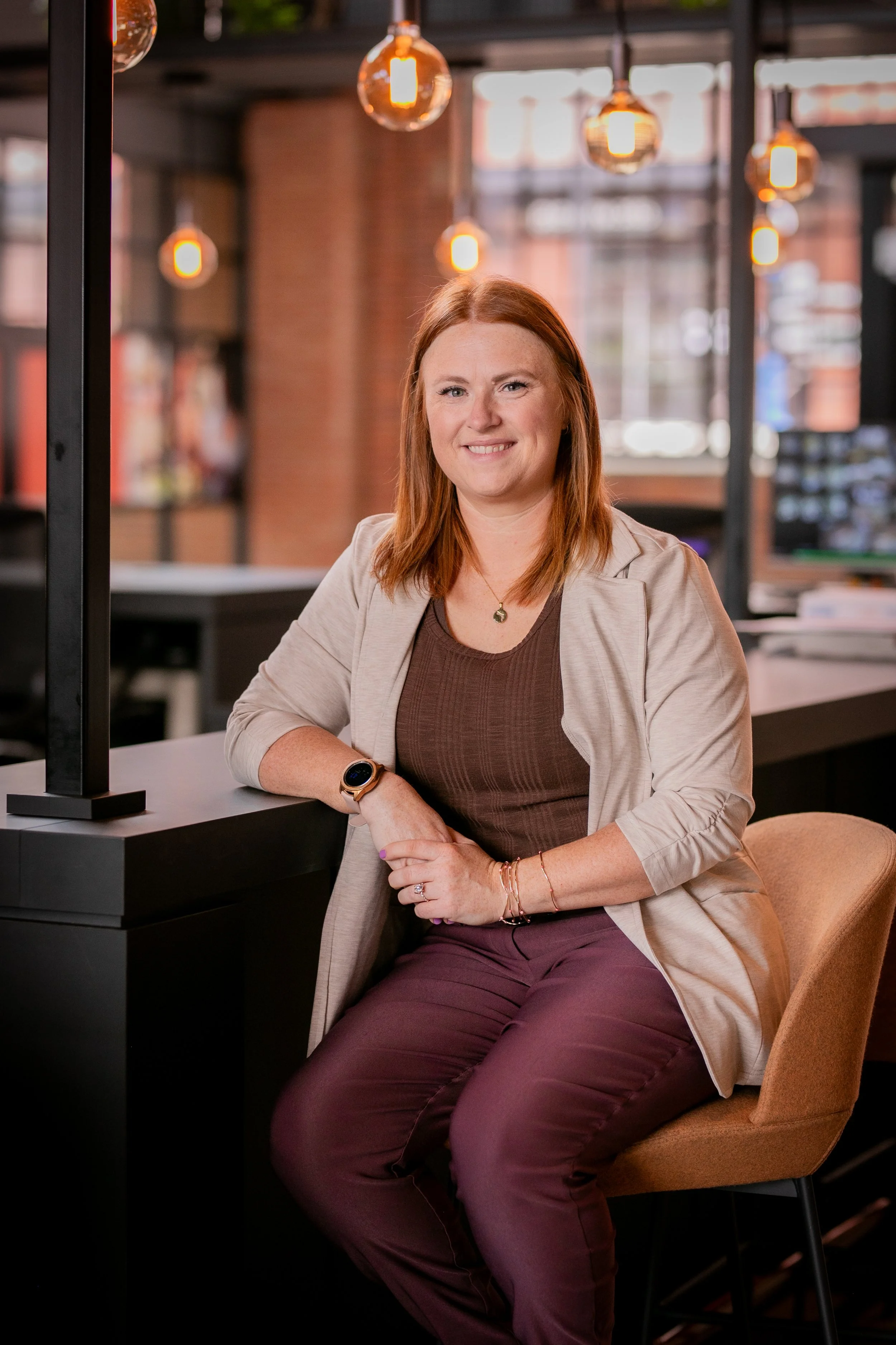 A woman with red hair sitting in a modern, well-lit indoor space with industrial decor, smiling at the camera, wearing a beige blazer, brown top, maroon pants, and a smartwatch.
