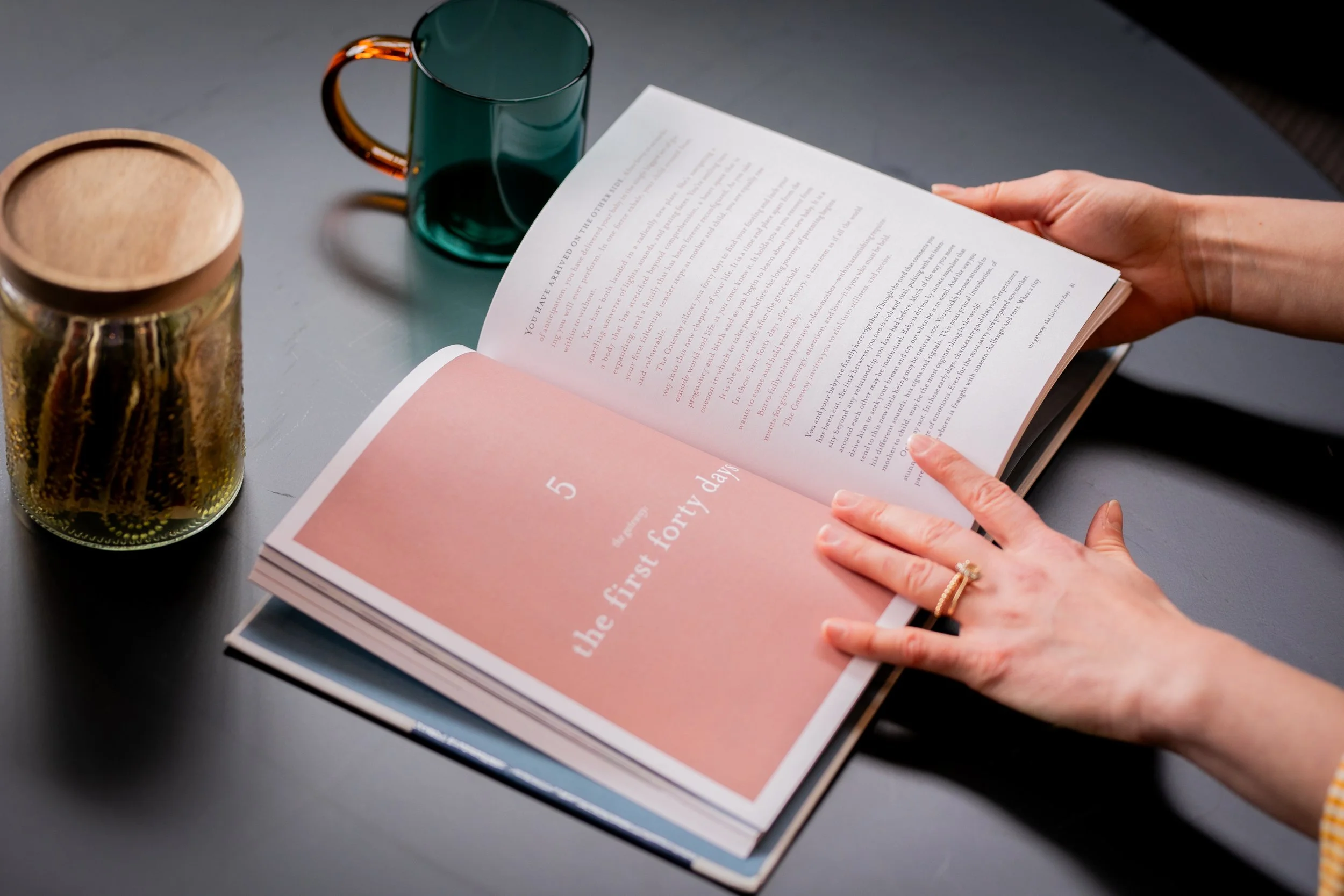 Person holding open a booklet titled "the first forty days" on a dark table, with a gold mug, a green glass mug, and a small container nearby.