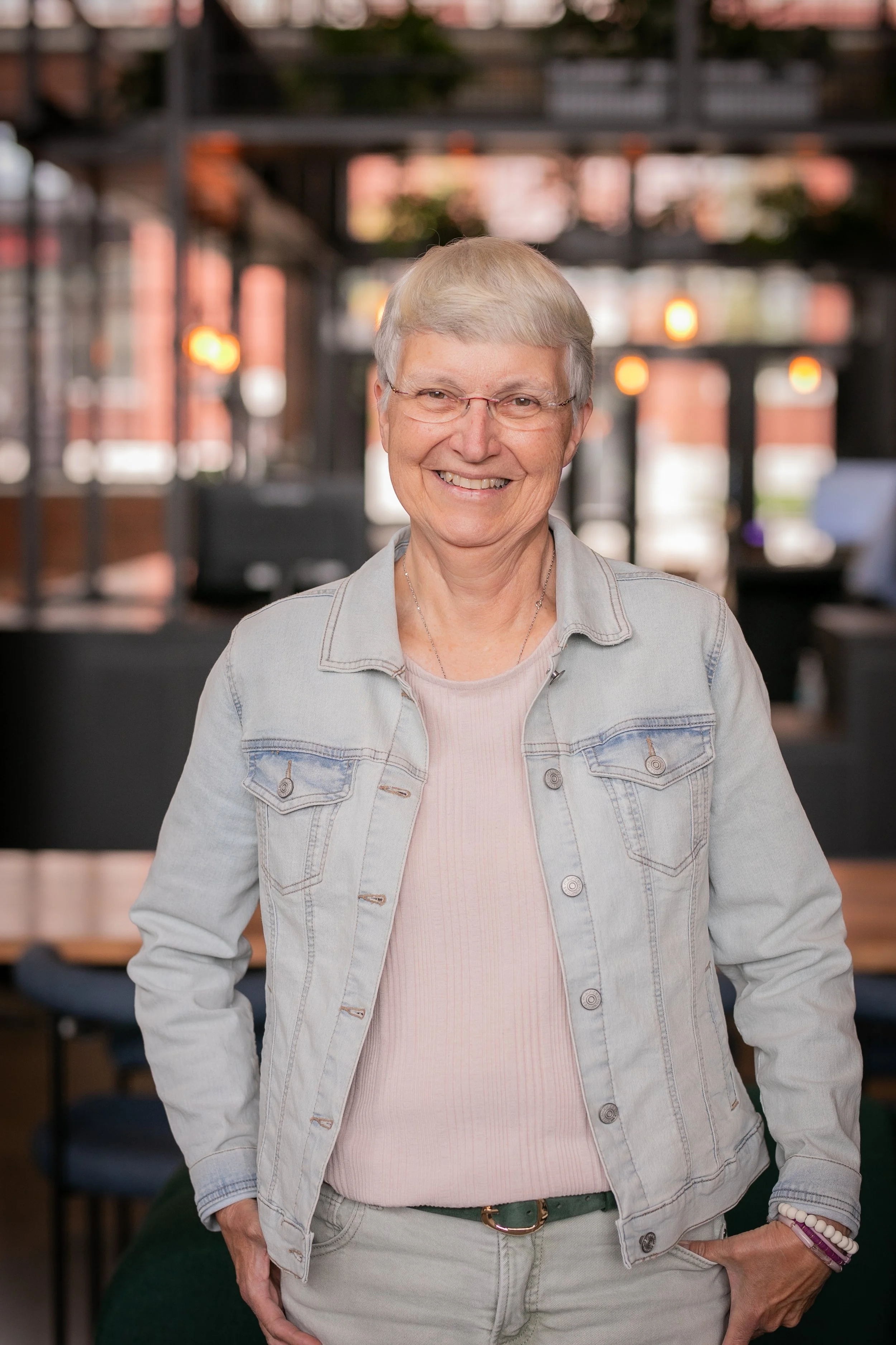 A smiling senior woman with short gray hair, glasses, wearing a light gray denim jacket over a pink top, standing in an indoor space with a blurred background of warm lights and dark furniture.