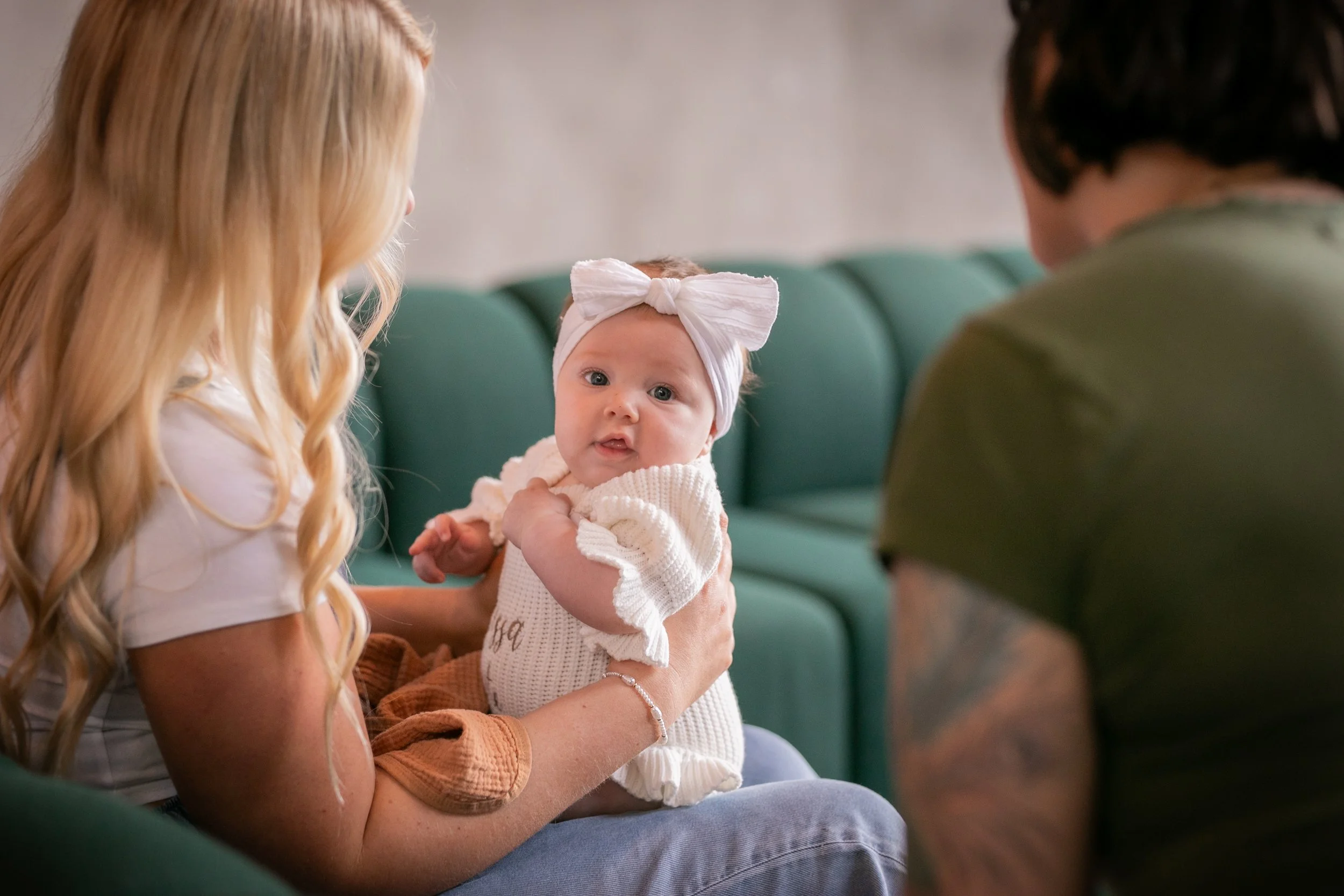A woman with long blonde hair holding a baby girl with a white bow headband, sitting on a person's lap in an indoor cafe or waiting area with a teal booth.