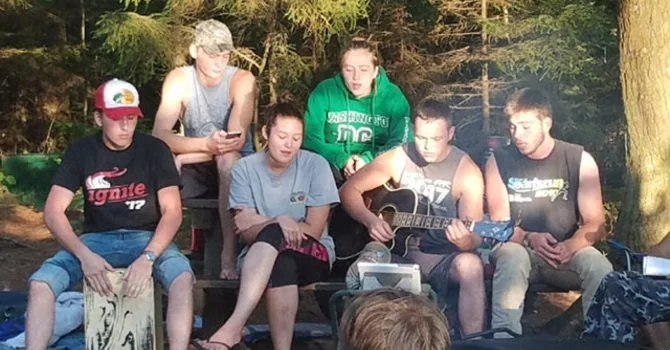 Six young people sitting outdoors around a campfire, with two women playing guitar, one person playing cajón, and others watching or using their phones.