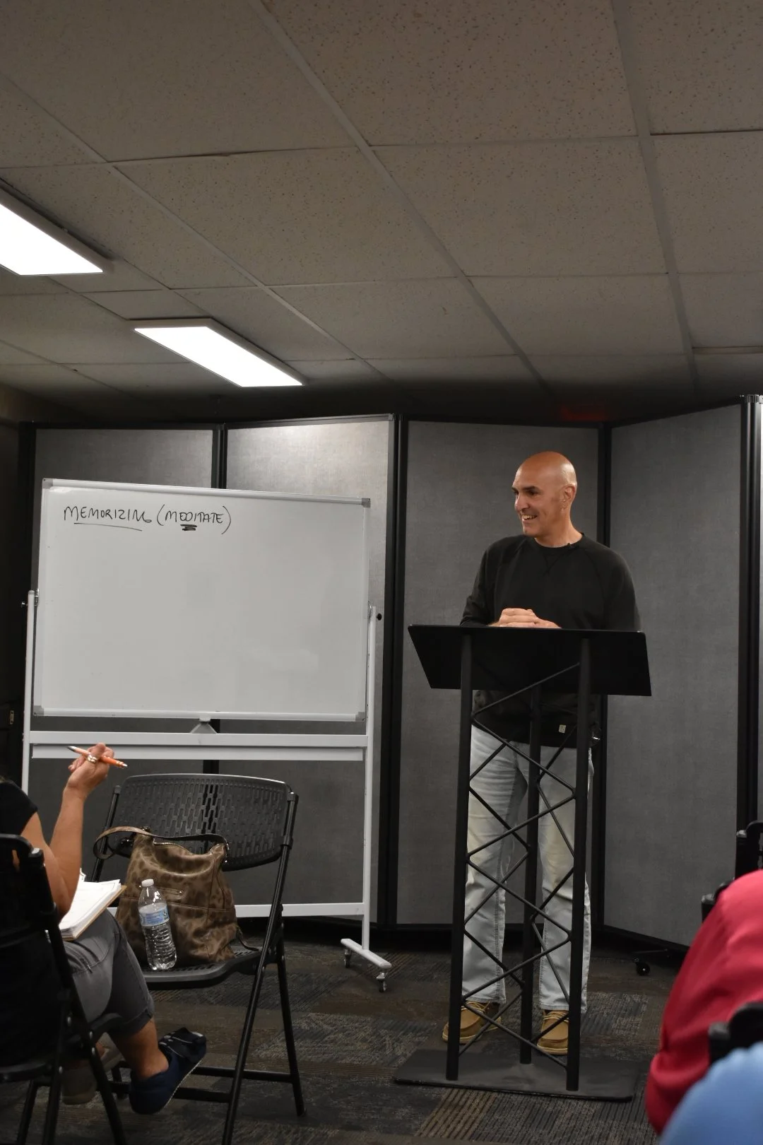 A man standing at a podium, smiling, in front of a whiteboard with the word 'MEMORIZING (MODIATE)' written on it, in a classroom or seminar room with gray partition walls and people seated.