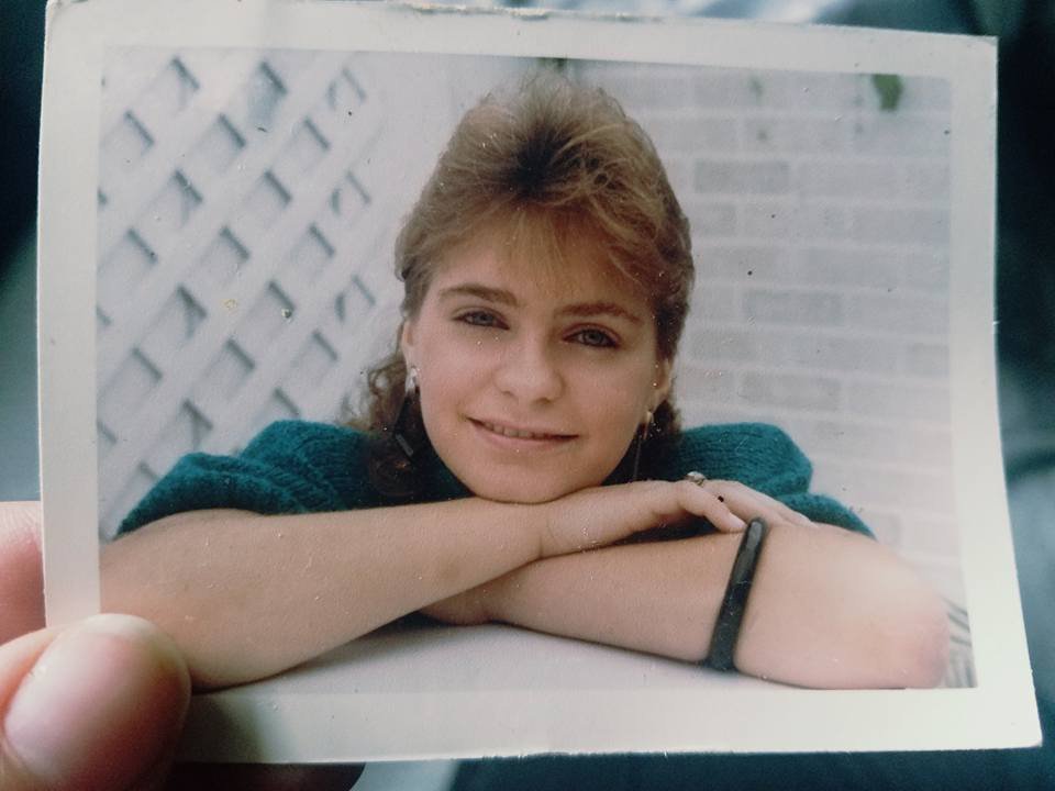 A woman with short, light brown hair, smiling and resting her chin on her crossed arms, wearing a teal sweater and earrings, standing outside in front of a white lattice and brick wall.
