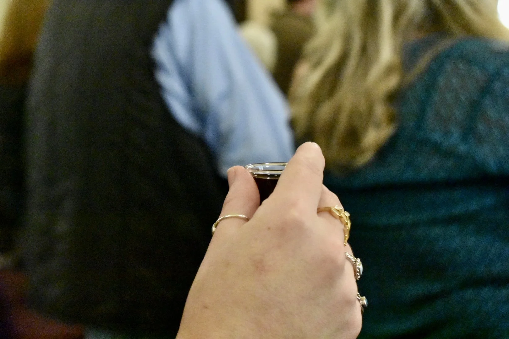 Close-up of a hand holding a shot glass with dark liquor, with bracelets and rings, in a social setting with people in the background.
