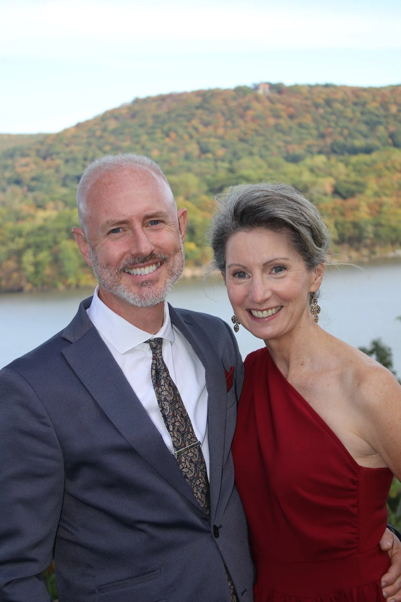 A smiling man in a suit and a smiling woman in a red dress standing outdoors with a river and a tree-covered hill in the background.